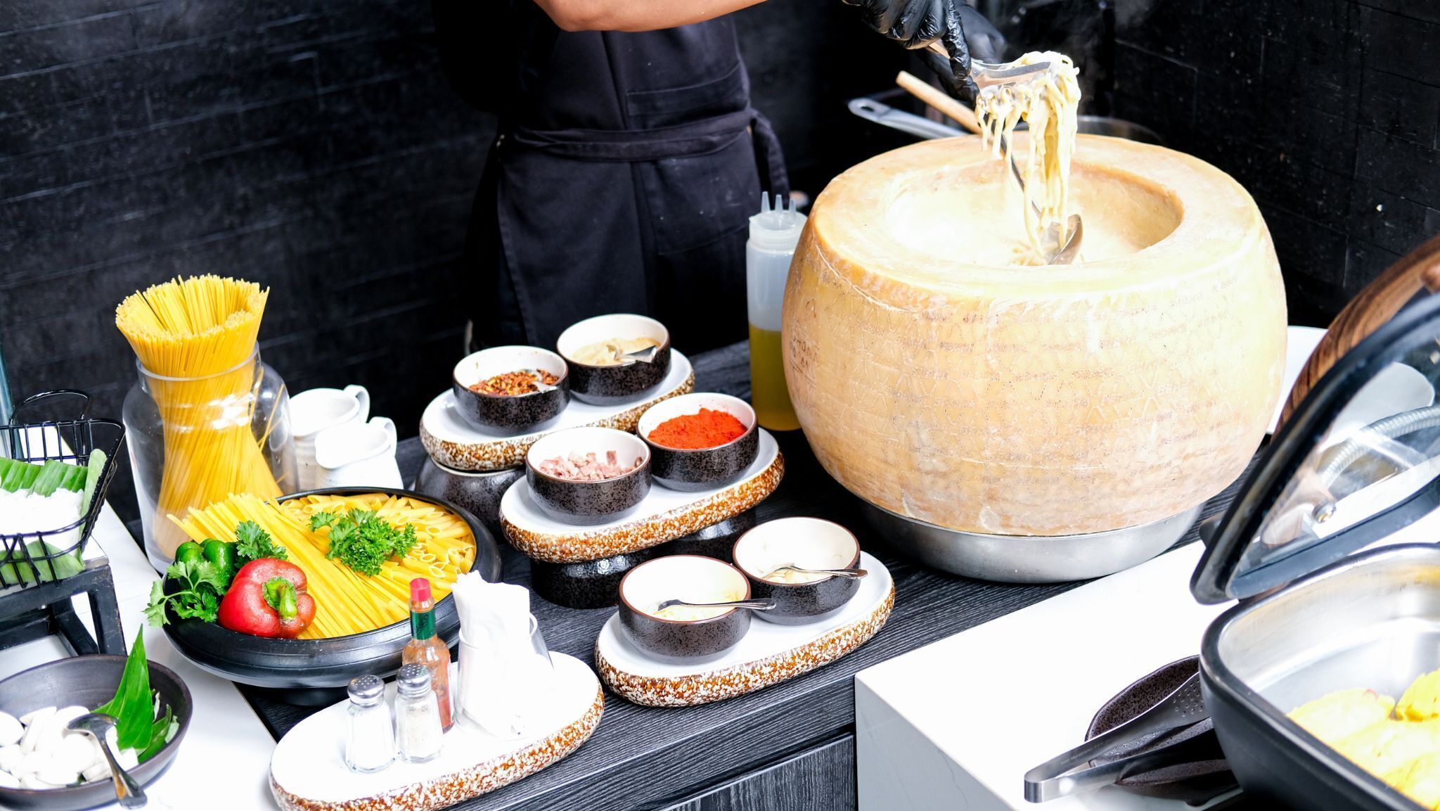 A buffet setup with pasta, various condiments, and a large cheese wheel.
