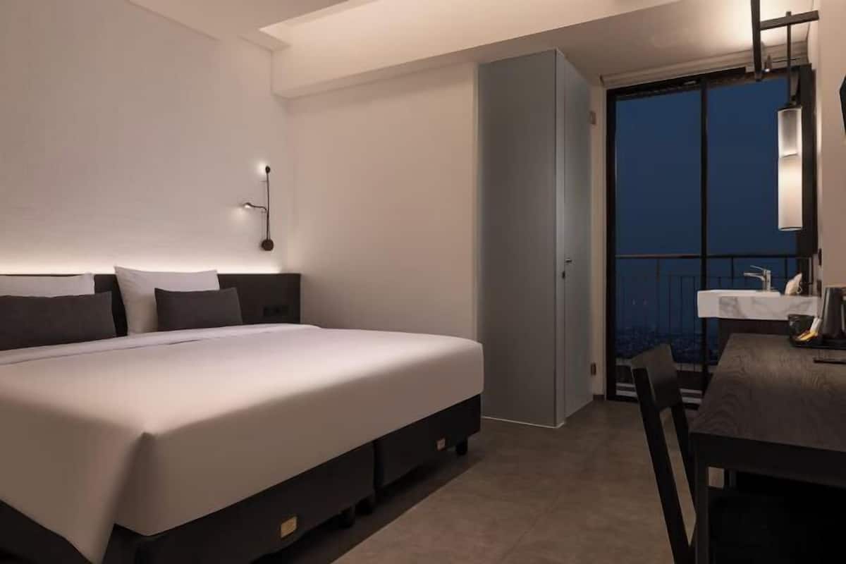 A modern hotel room with a large bed, a small table, a nightstand, and a balcony with a view.
