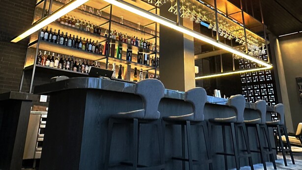 Zona de bar con estantes llenos de botellas de licor, una barra y taburetes.
