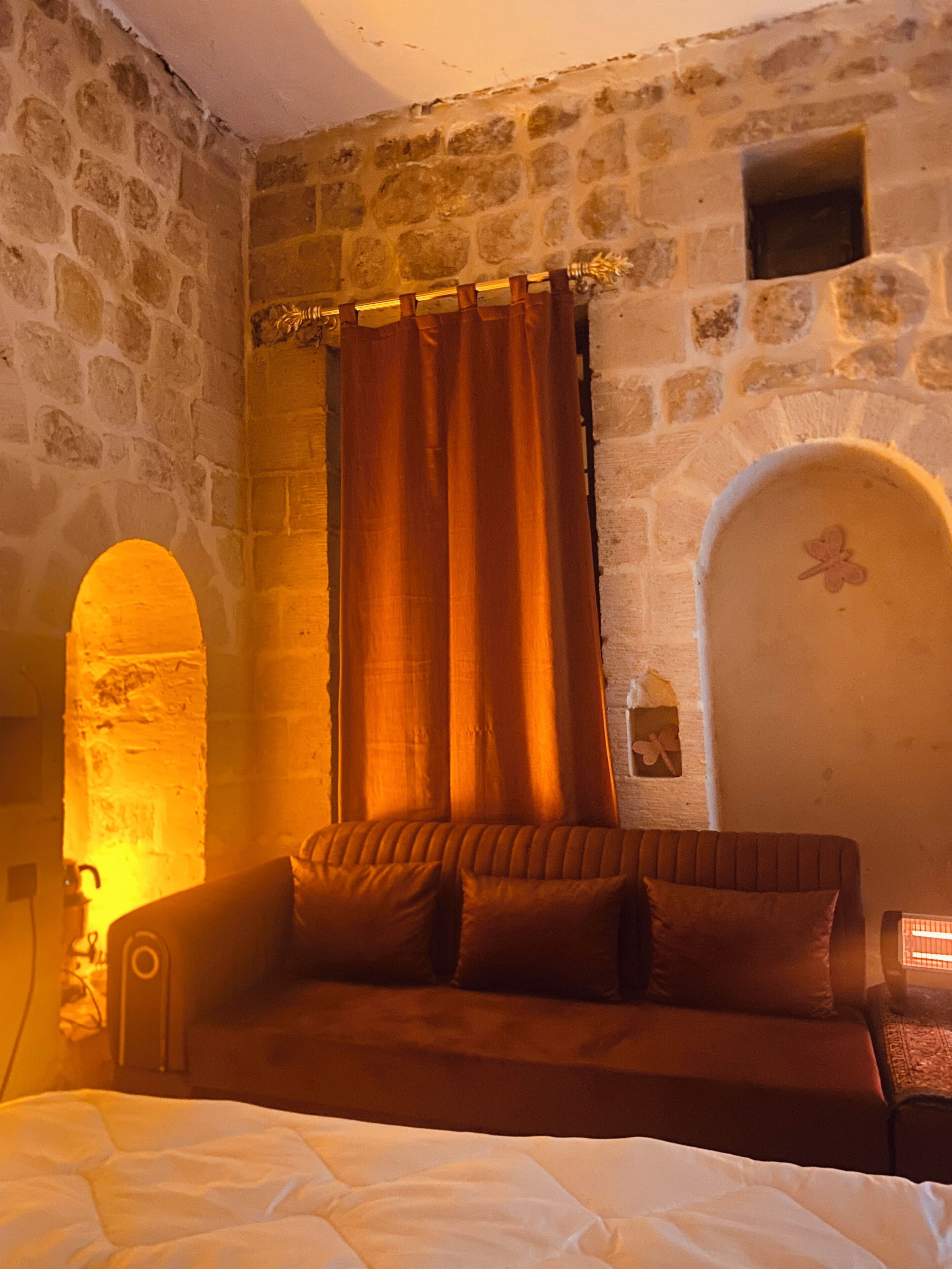 Una habitación con pared de piedra, cama con ropa de cama blanca, sofá marrón con cojines y una cortina.
