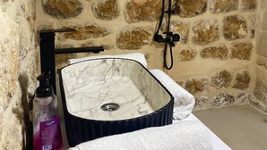 A bathroom with a stone wall, a marble sink, a handheld showerhead, a bottle of shampoo, and a towel. - Roma Konağı Mardin (Mardin)