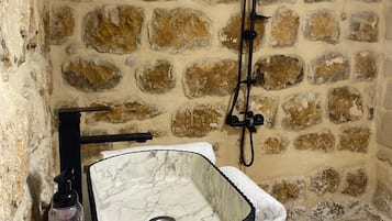 A bathroom with a stone wall, a marble sink, a handheld showerhead, a bottle of shampoo, and a towel.
