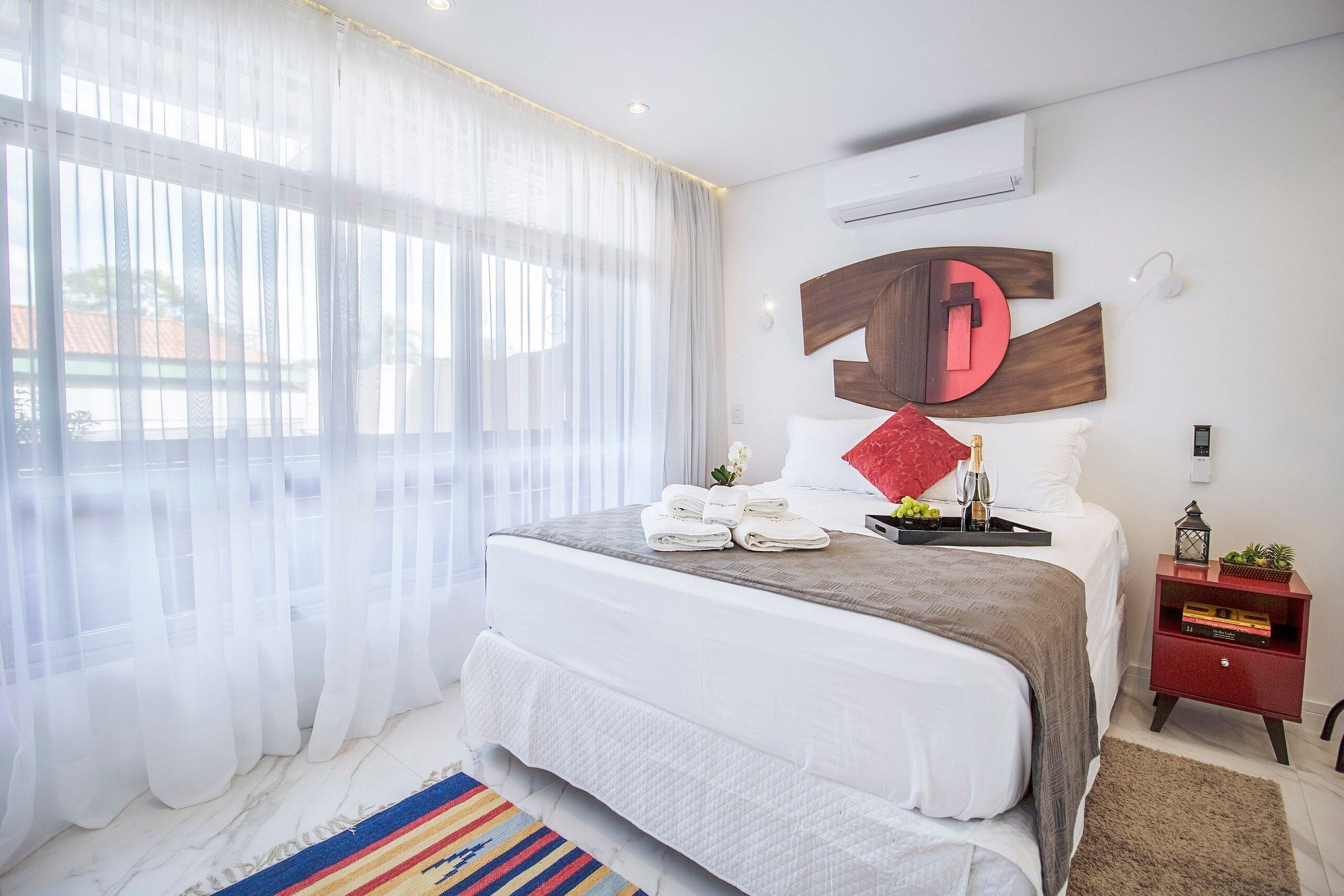 Ein modernes Schlafzimmer mit einem großen Bett, weißer Bettwäsche, einem roten Zierkissen, einem Nachttisch mit roter Lampe und einer wandmontierten Klimaanlage.