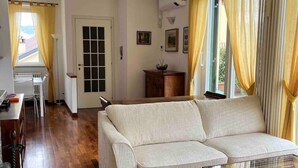 A living room with a sofa, wooden flooring, a glass coffee table, and a view of the outdoors through a window with curtains. - The Terrace on the lake (Fiumelatte)