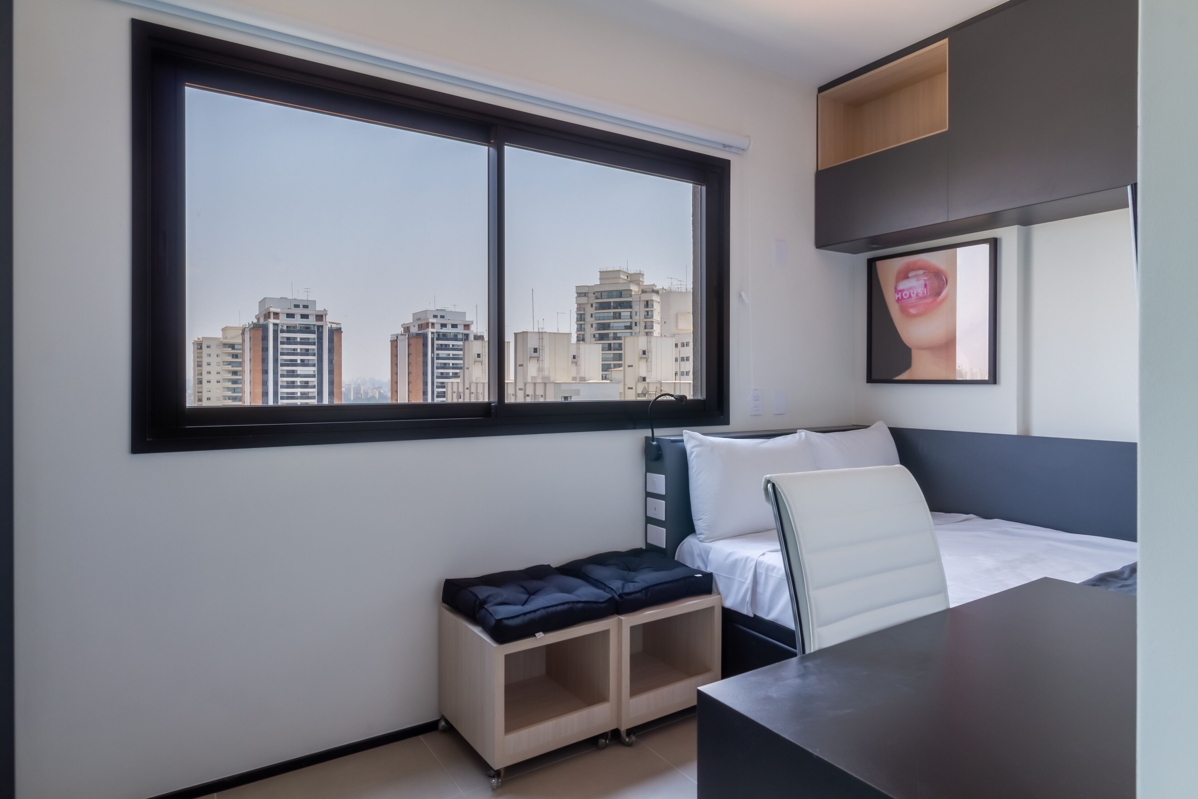 A modern hotel room with a large window, a bed with white bedding, a desk, and a small seating area.