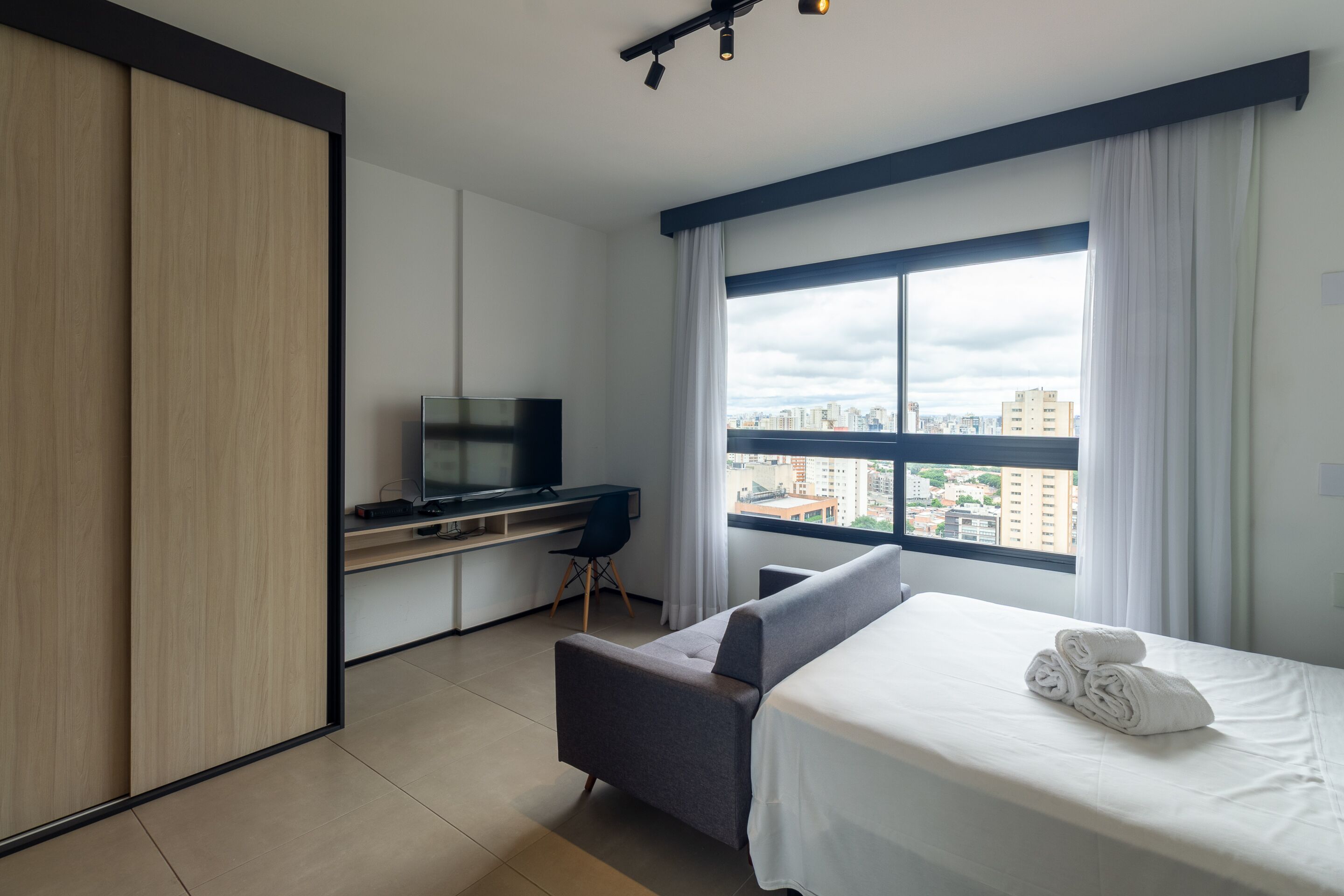 Ein modernes Hotelzimmer mit Bett, Schreibtisch mit Fernseher, Blick auf die Stadt und einem großen Fenster.