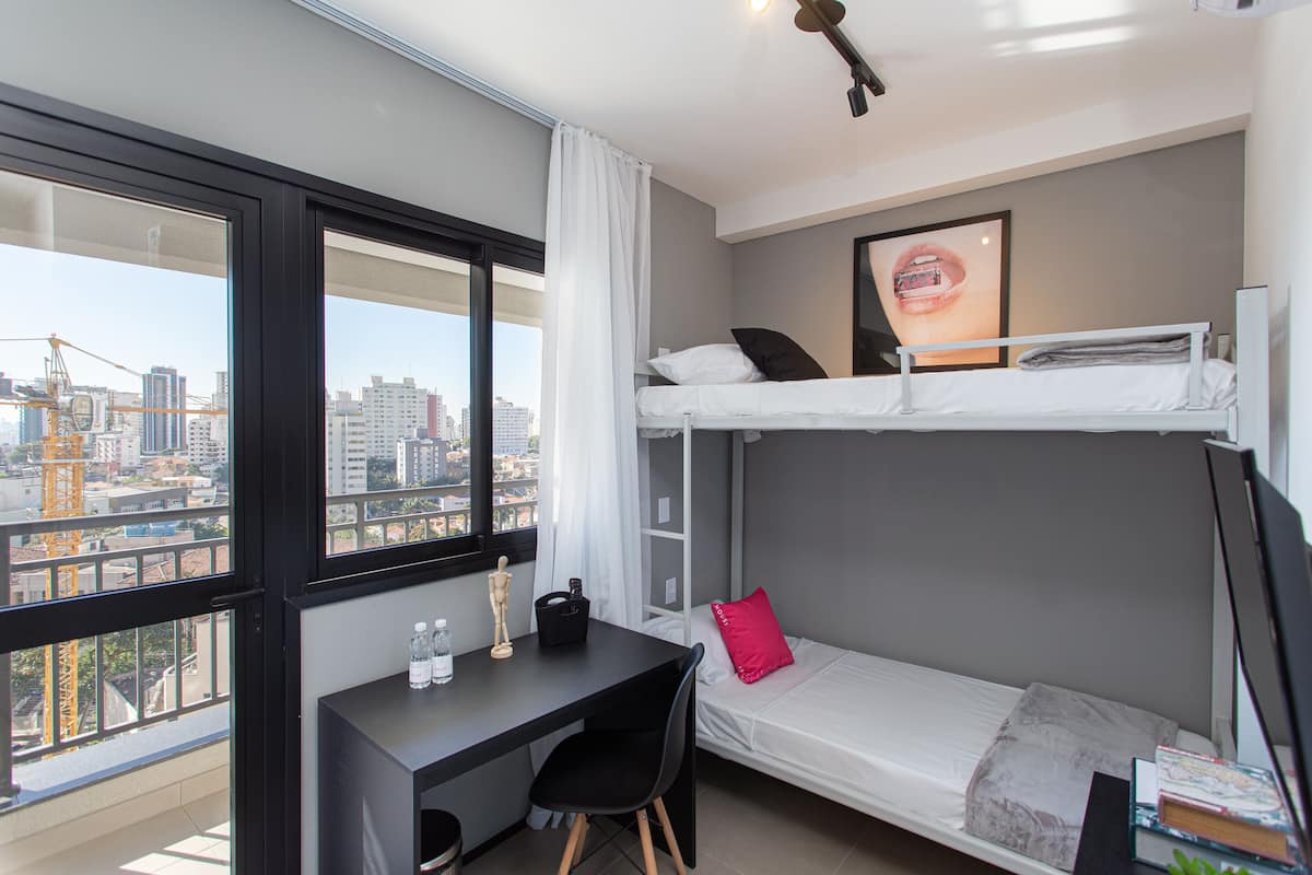 Een kleine hotelkamer met een stapelbed, een bureau met stoel, een tafeltje en een raam met uitzicht over de stad.
