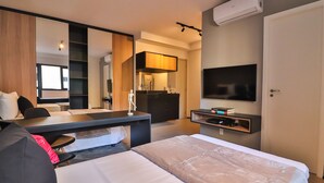 A modern hotel room with a large bed, a desk with a chair, a flat-screen TV mounted on the wall, a built-in kitchen area, and a large mirror. - 360 VN Humberto (São Paulo)