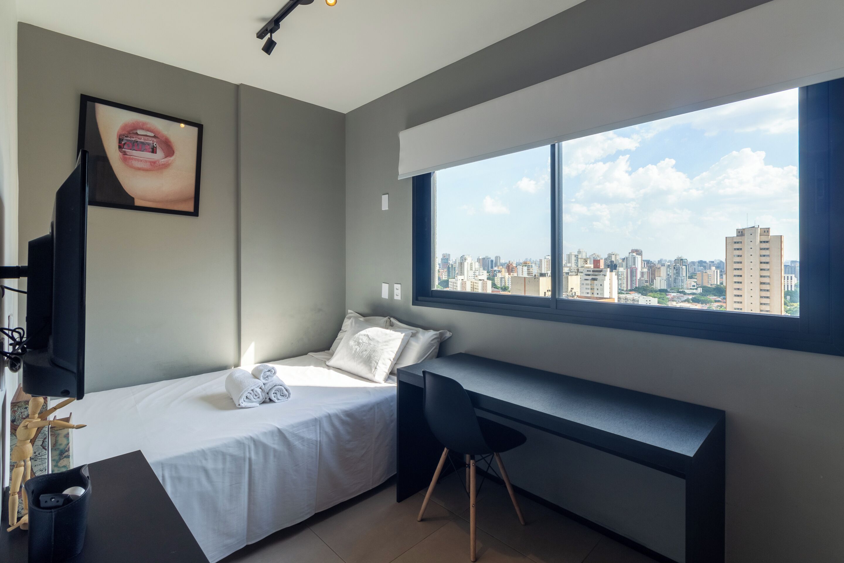 A modern bedroom with a bed, a desk, and a large window offering a city view.