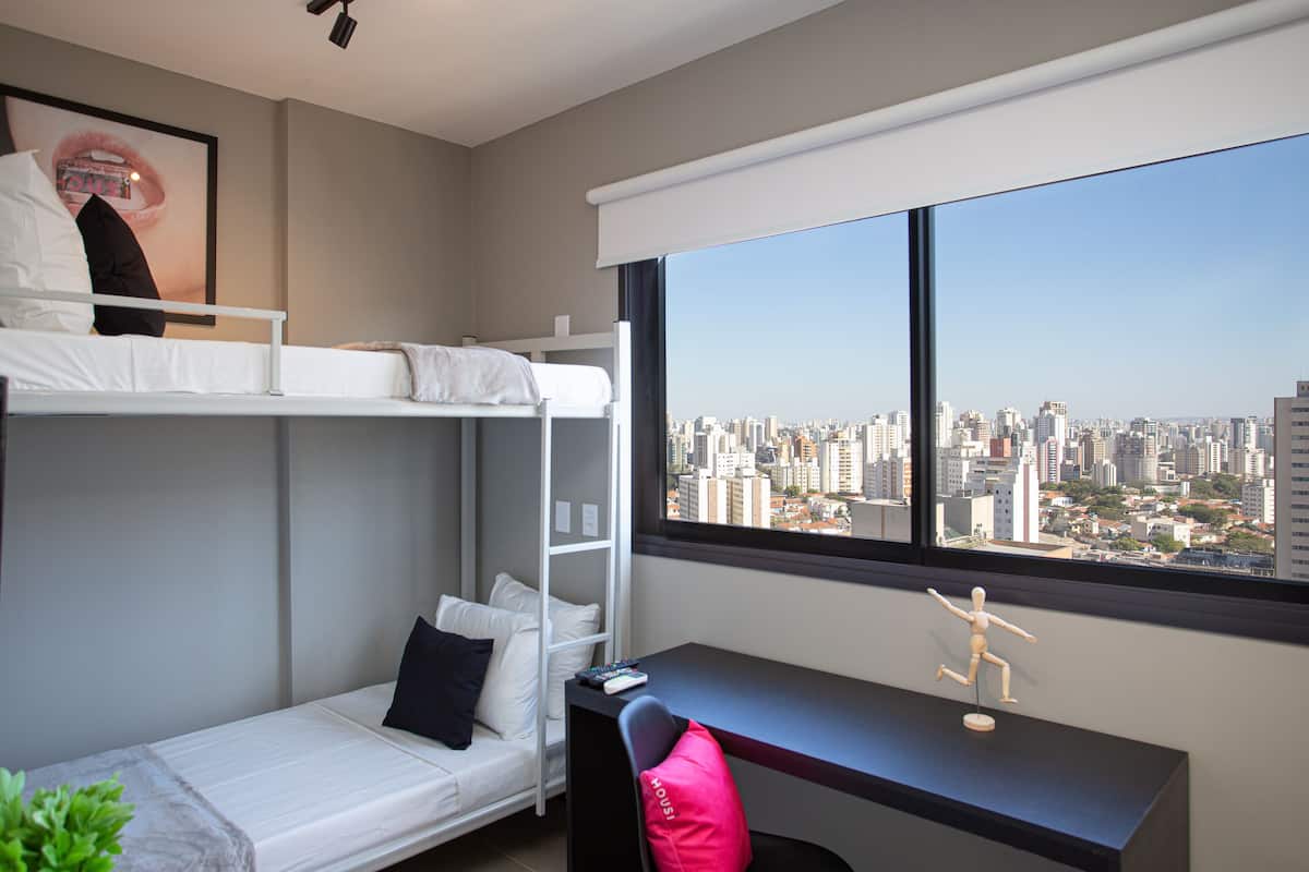 Een hotelkamer met een stapelbed, een bureau met een stoel, een groot raam met uitzicht op de stad en een kleine plant op de vensterbank.