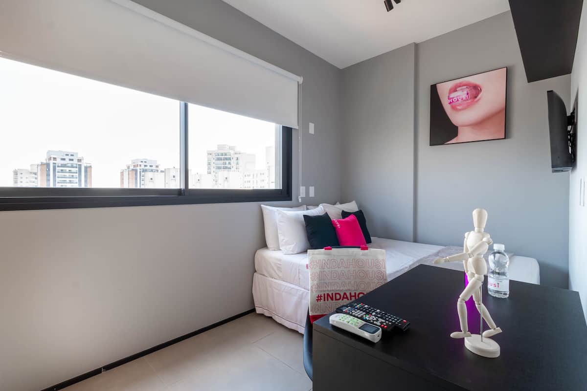 Een moderne hotelkamer met een groot raam, een bed met wit beddengoed en kleurrijke kussens, een bureau met een etalagepop, een waterfles en een afstandsbediening.