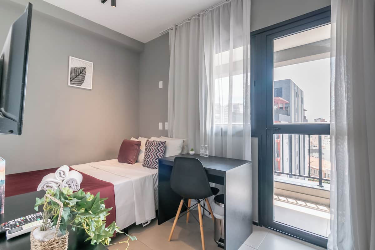 Een moderne hotelkamer met een bed, bureau en stoel, een kleine plant en uitzicht op de stad door een groot raam.