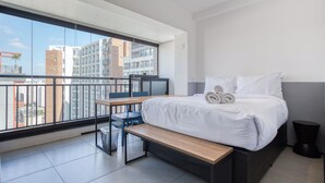 A modern hotel room with a large balcony, a bed with white linens, a wooden bench, and a small table. - 360 VN Humberto (São Paulo)