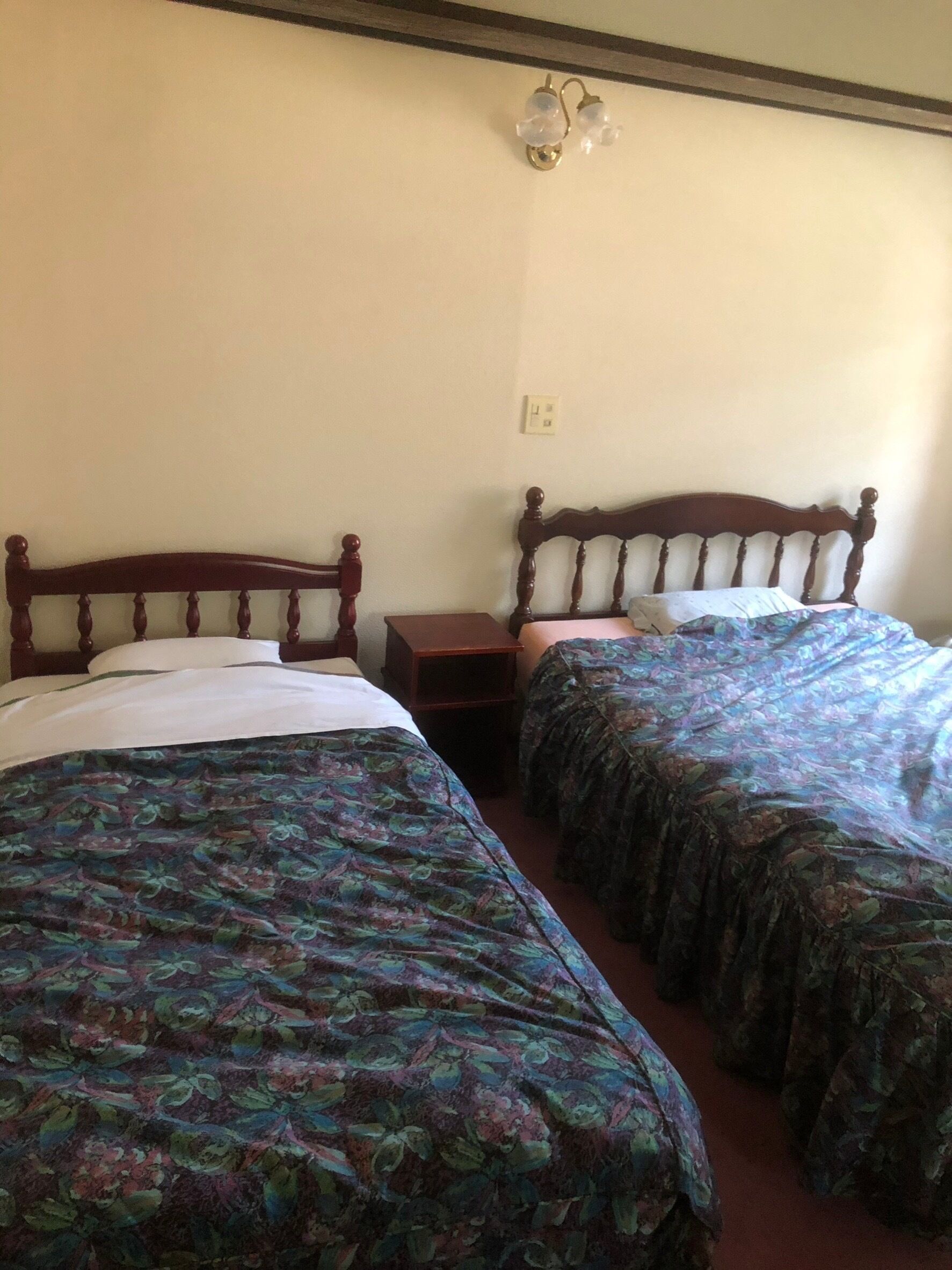 A room with two single beds with floral-patterned bedding, a wooden headboard, a nightstand, and a wall-mounted light fixture.