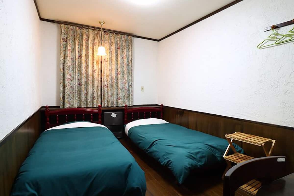 A small, compact room with two single beds, a wooden bench, a small table, and a hanging clothes rack.