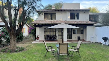 A two-story house with a patio area featuring a table and four chairs, a large umbrella, and a barbecue grill.