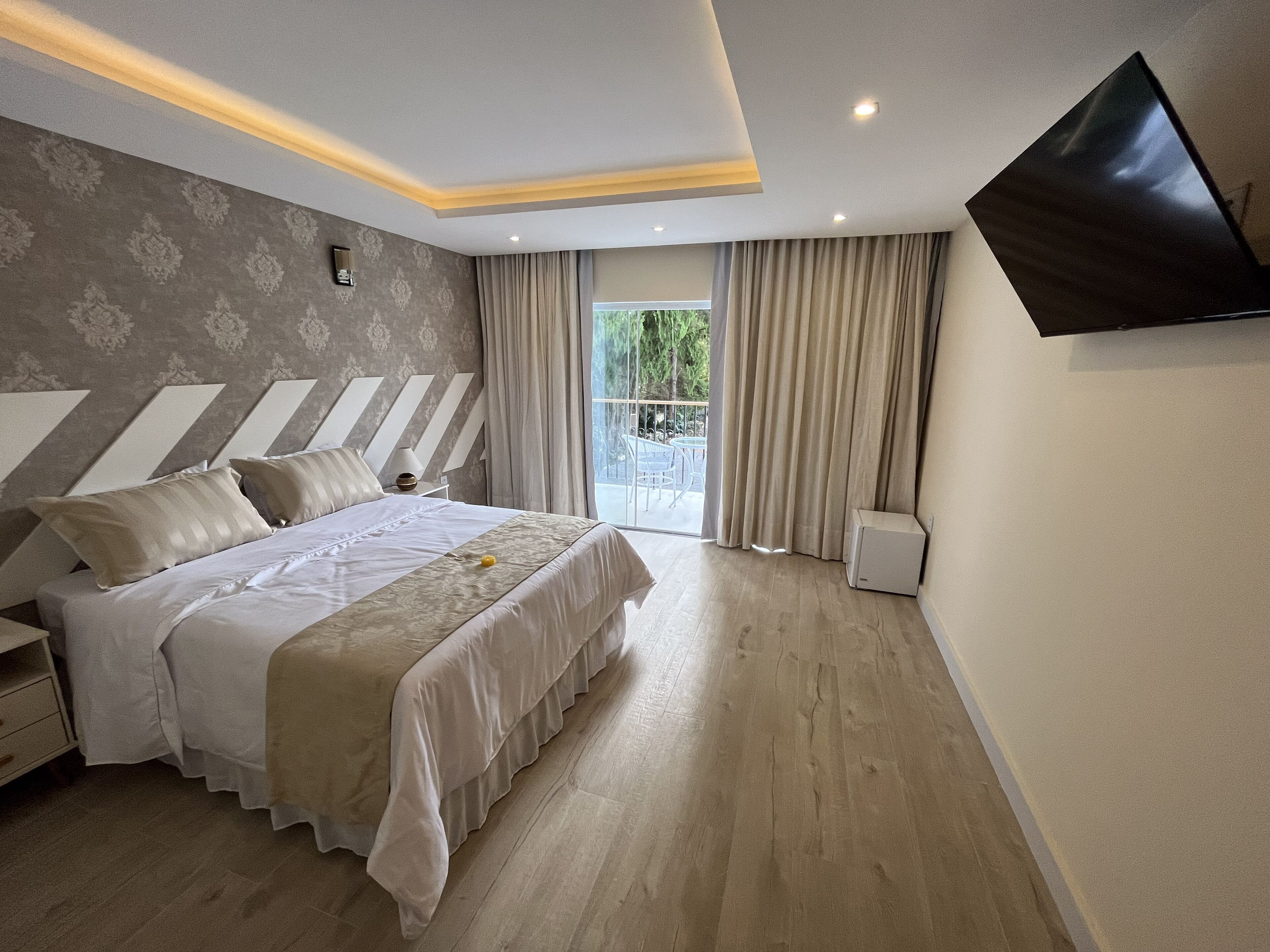 A modern bedroom with a large bed, a flat-screen TV mounted on the wall, and a balcony with curtains.