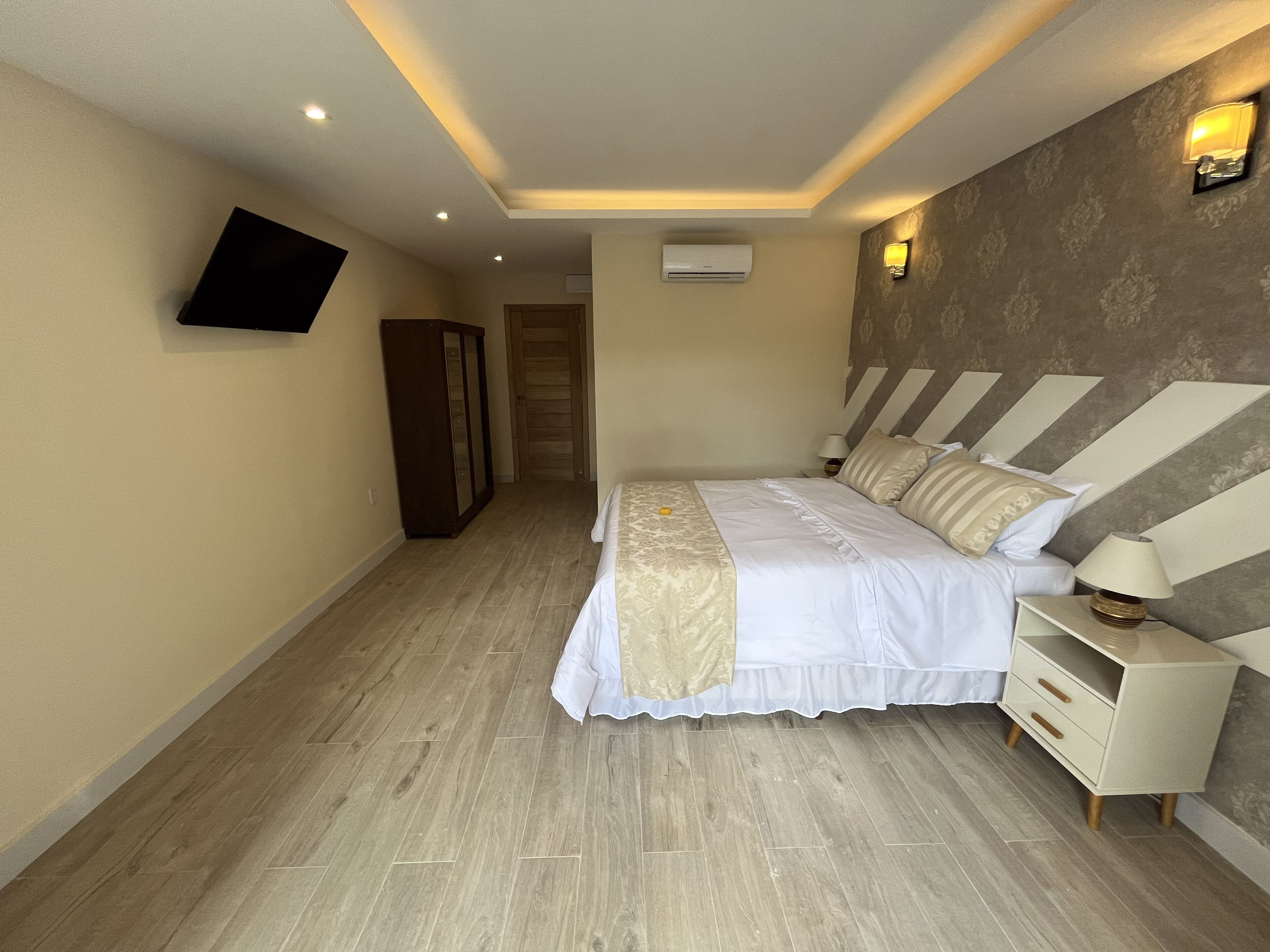 A bedroom with a bed, bedside table, lamp, wall-mounted TV, wardrobe, and an air conditioning unit.
