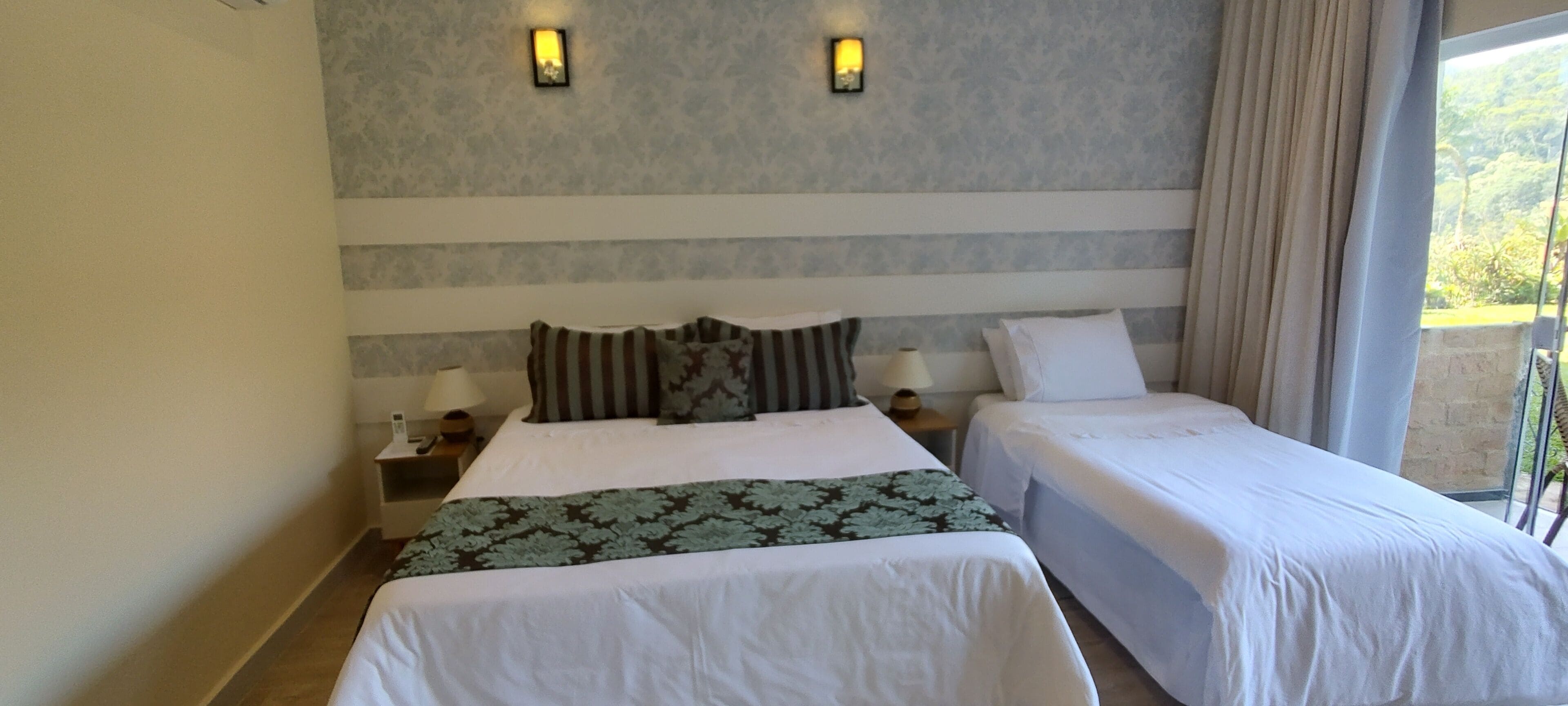 A hotel room with two beds, a nightstand with lamps, a wall-mounted light fixture, and a view of greenery outside.