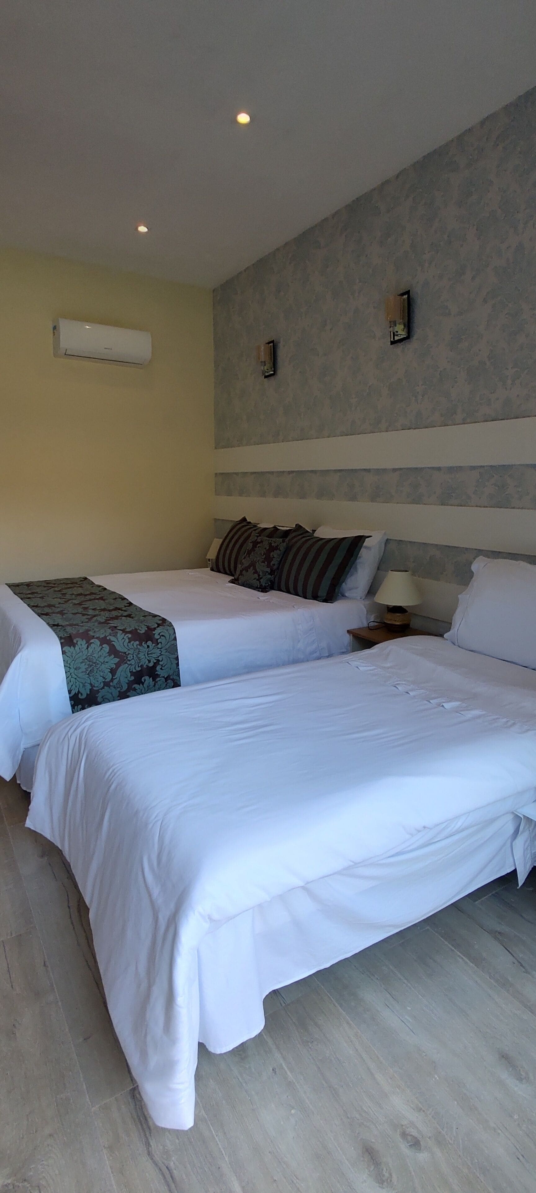 A hotel room with two single beds, a small bedside table with a lamp, a wall-mounted air conditioner, and a patterned wall.