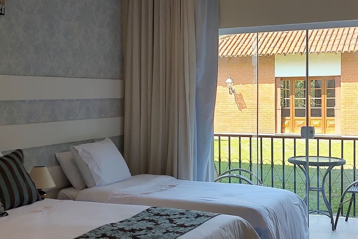 A hotel room with two beds, a balcony with a view of a building, and a window with curtains.