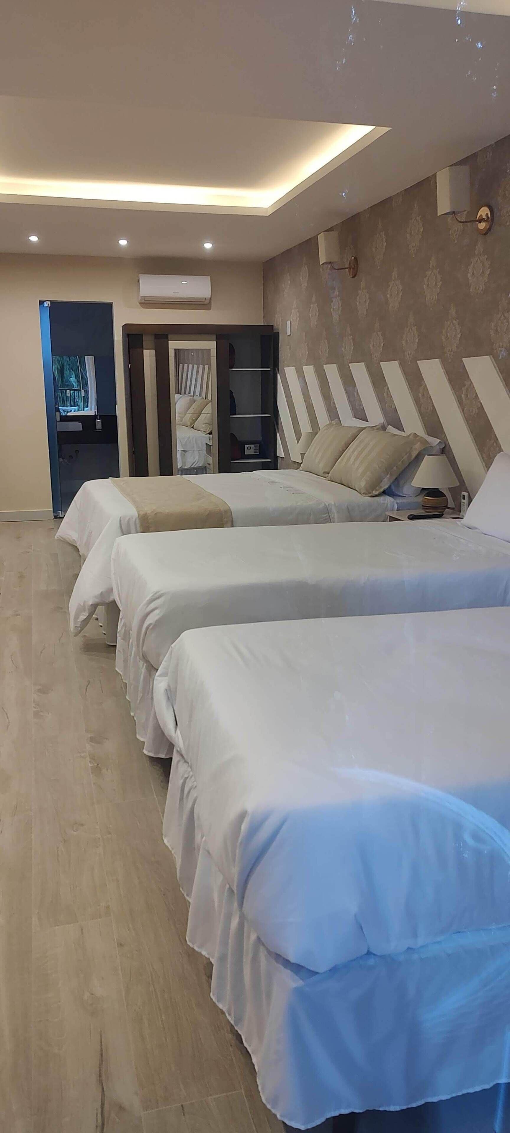 Quarto de hotel com três camas, um guarda-roupa, uma televisão e ar condicionado.