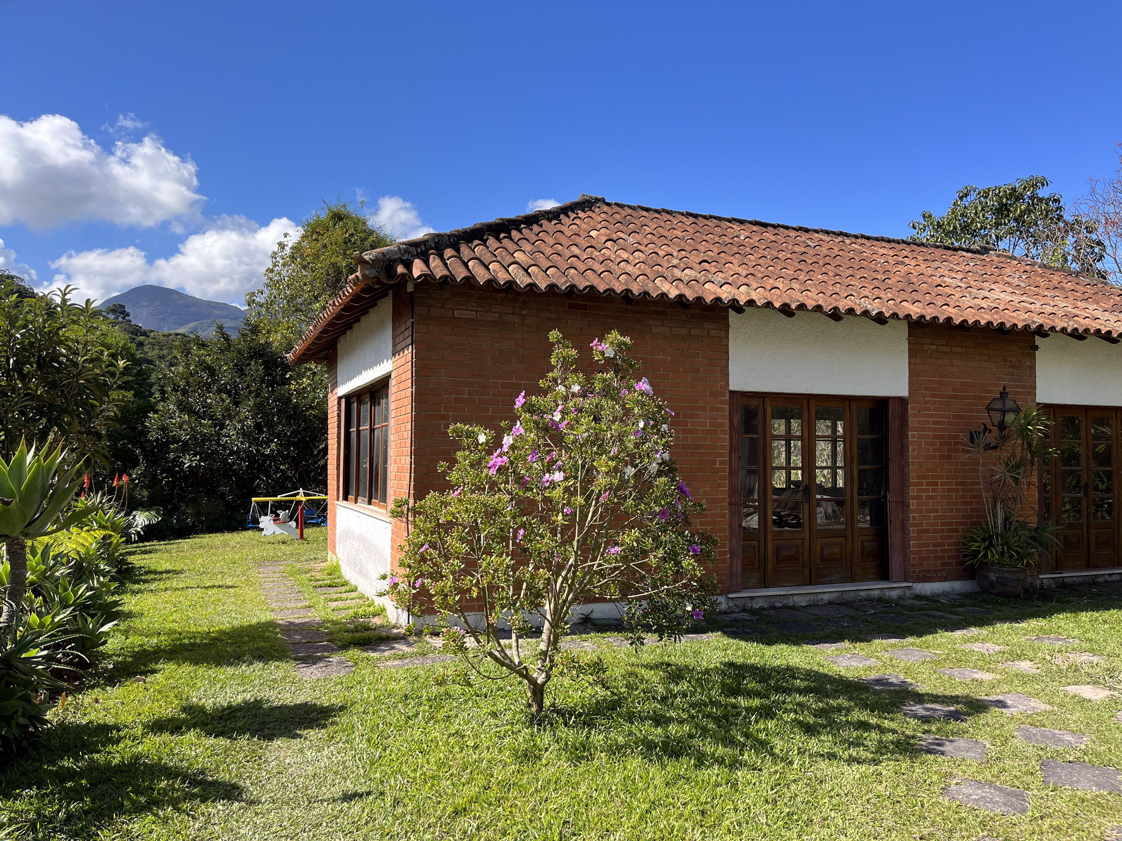 A single-story brick house with a tiled roof, a small front yard with a tree, and a mountain in the background.