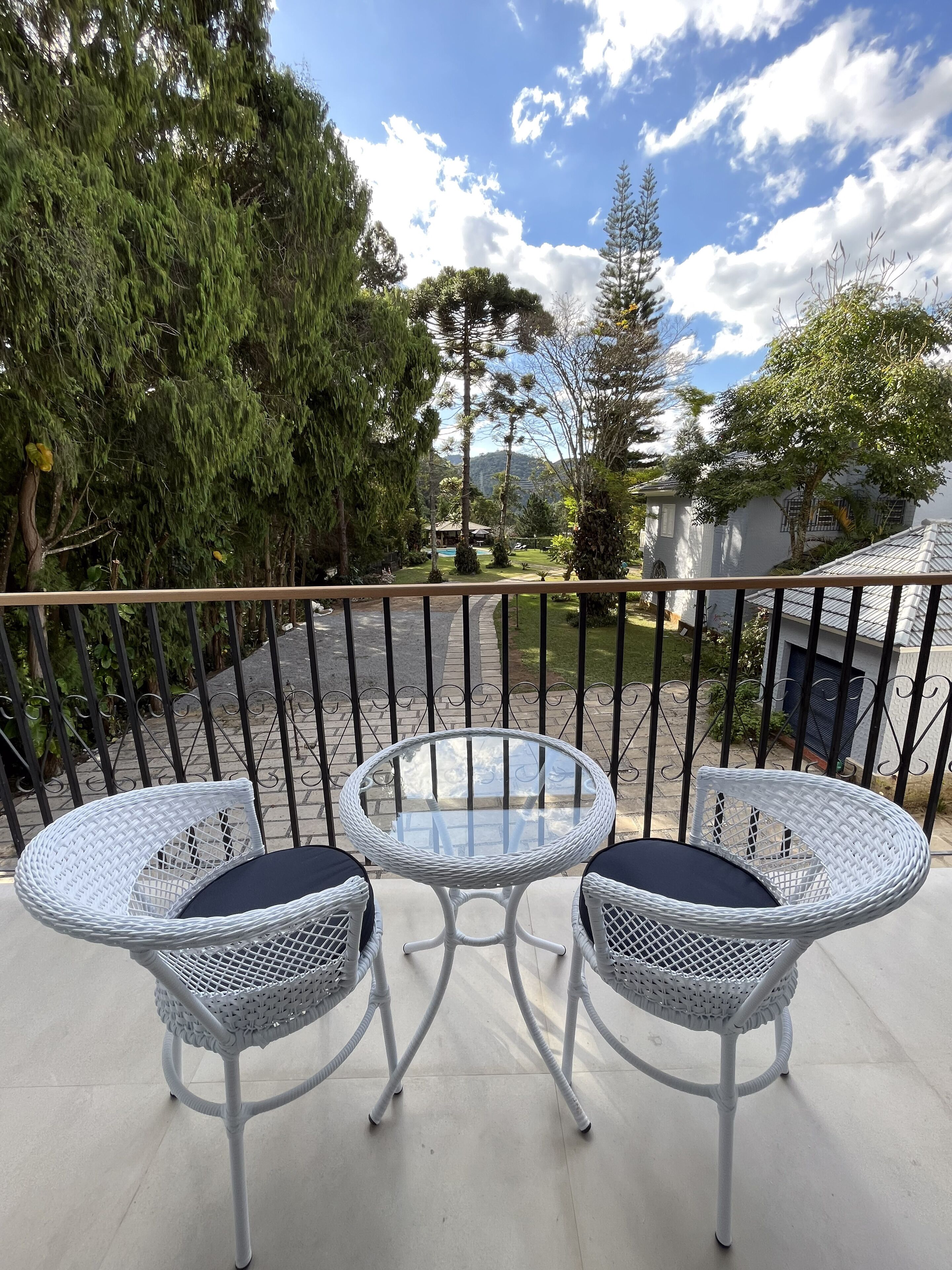 A balcony with two white wicker chairs and a glass table, overlooking a garden with trees and a building in the background.