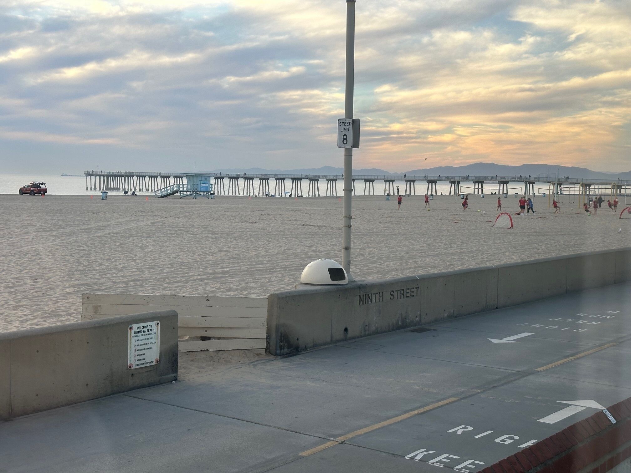 En strand med en fartgrænskeplade, en bro i baggrunden og en velkomstskilt til Hermosa Beach.