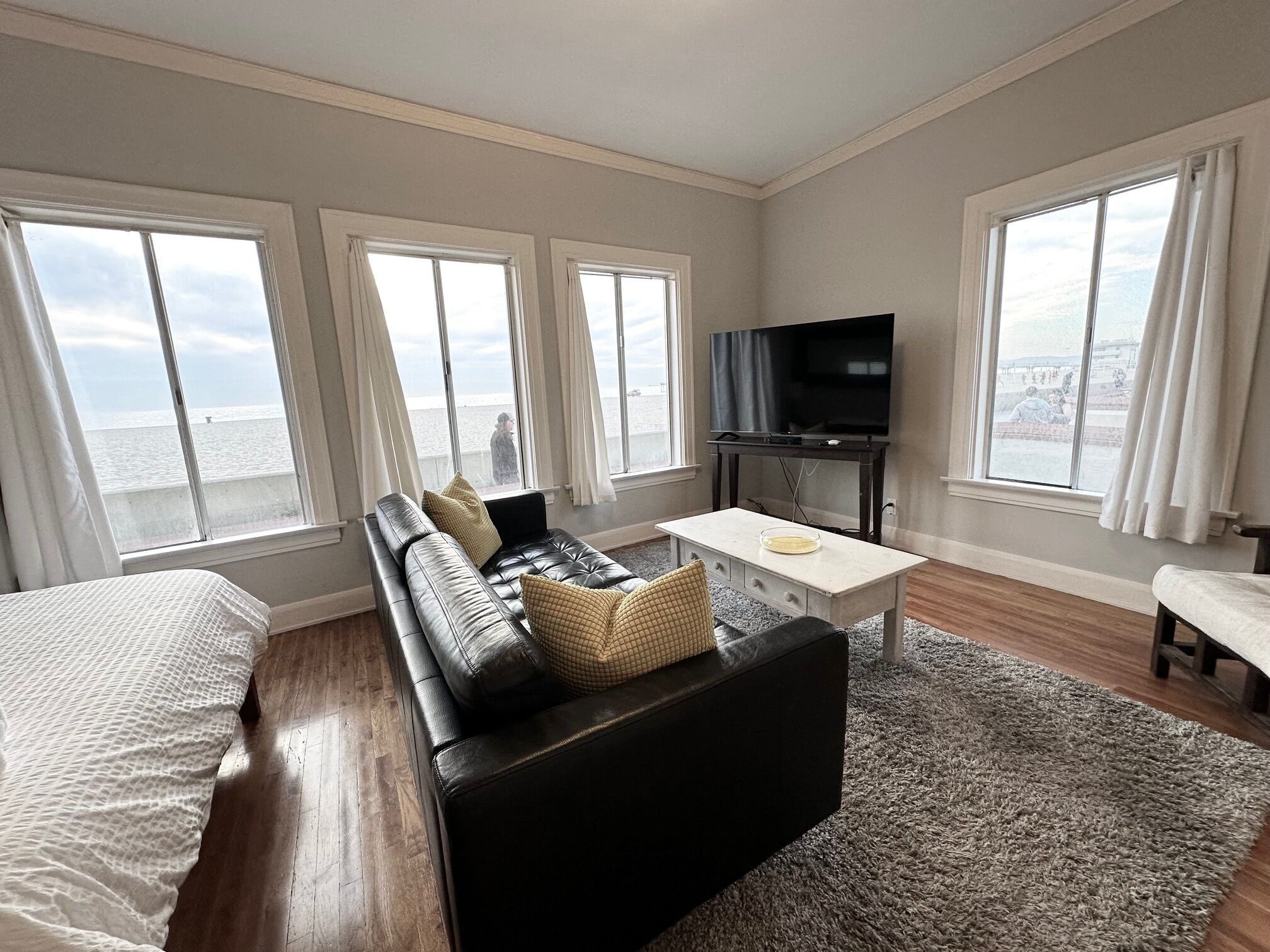 Una sala de estar moderna con un sofá de cuero negro, una mesita blanca y un televisor sobre un mueble bajo. Amplias ventanas ofrecen vistas a la ciudad.
