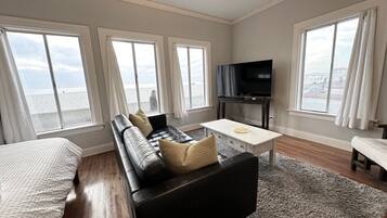 Una sala de estar moderna con un sofá de cuero negro, una mesita blanca y un televisor sobre un mueble bajo. Amplias ventanas ofrecen vistas a la ciudad.