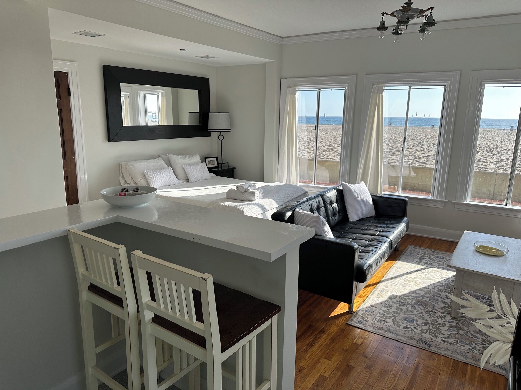 A bedroom with a bed, a black leather sofa, a small table, a white chair, and a view of the beach.