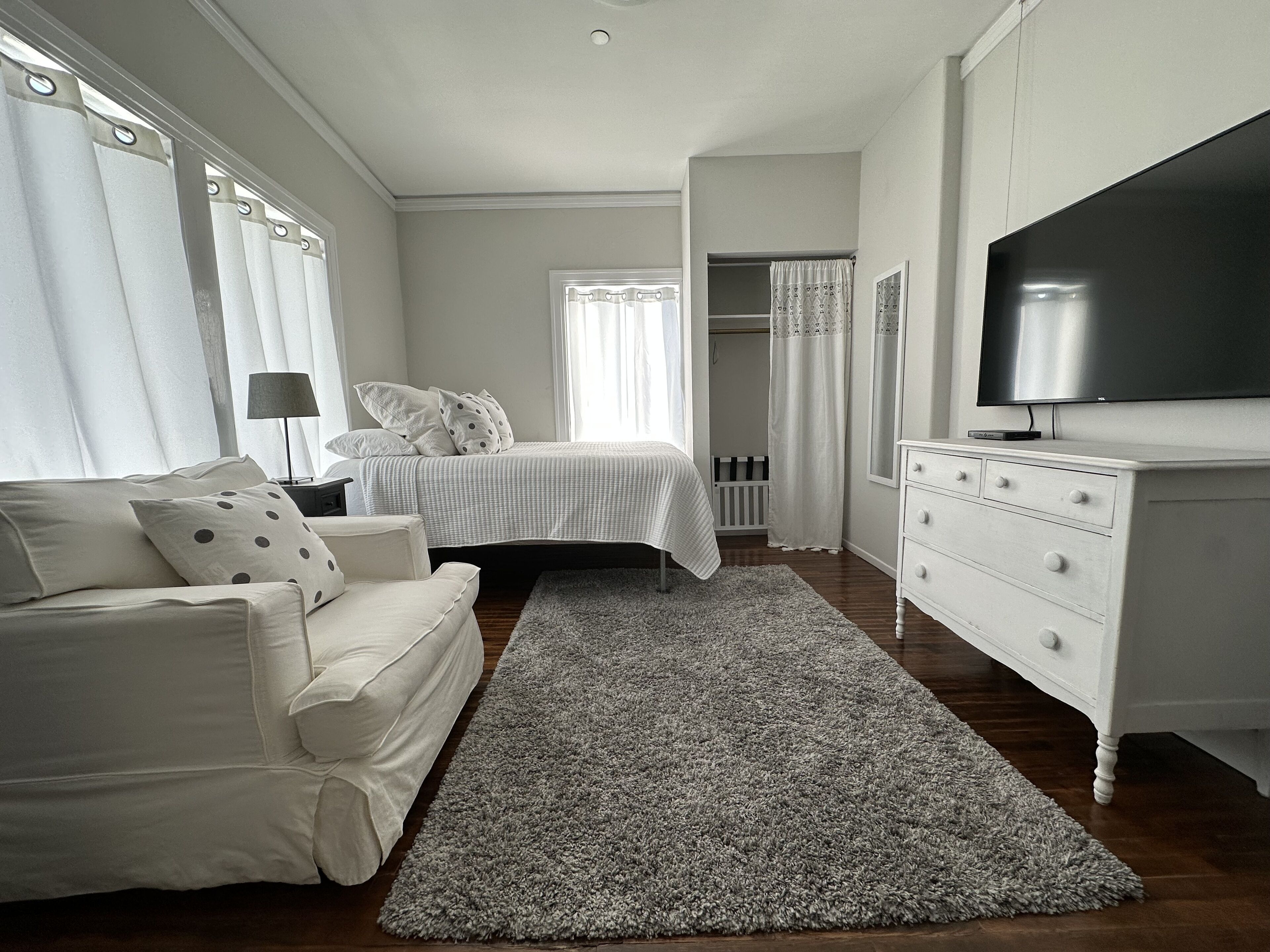 Quarto com cama, penteadeira branca, televisão e janela com cortinas.