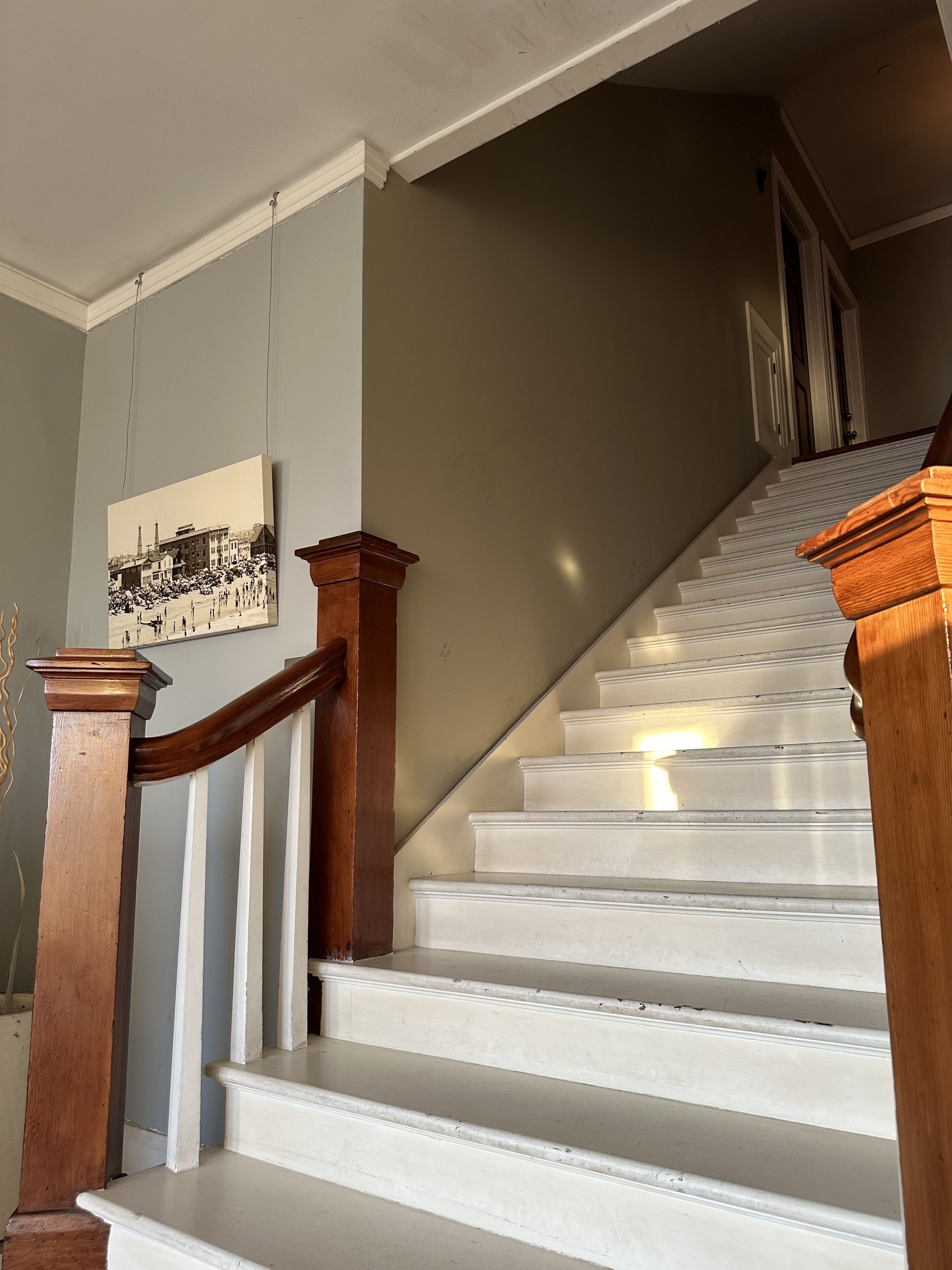 Un escalier avec des marches blanches et des rampes en bois, menant à un palier où est accrochée une image encadrée.