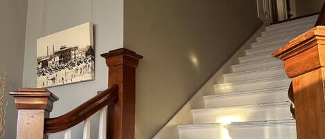 Un escalier avec des marches blanches et des rampes en bois, menant à un palier où est accrochée une image encadrée.