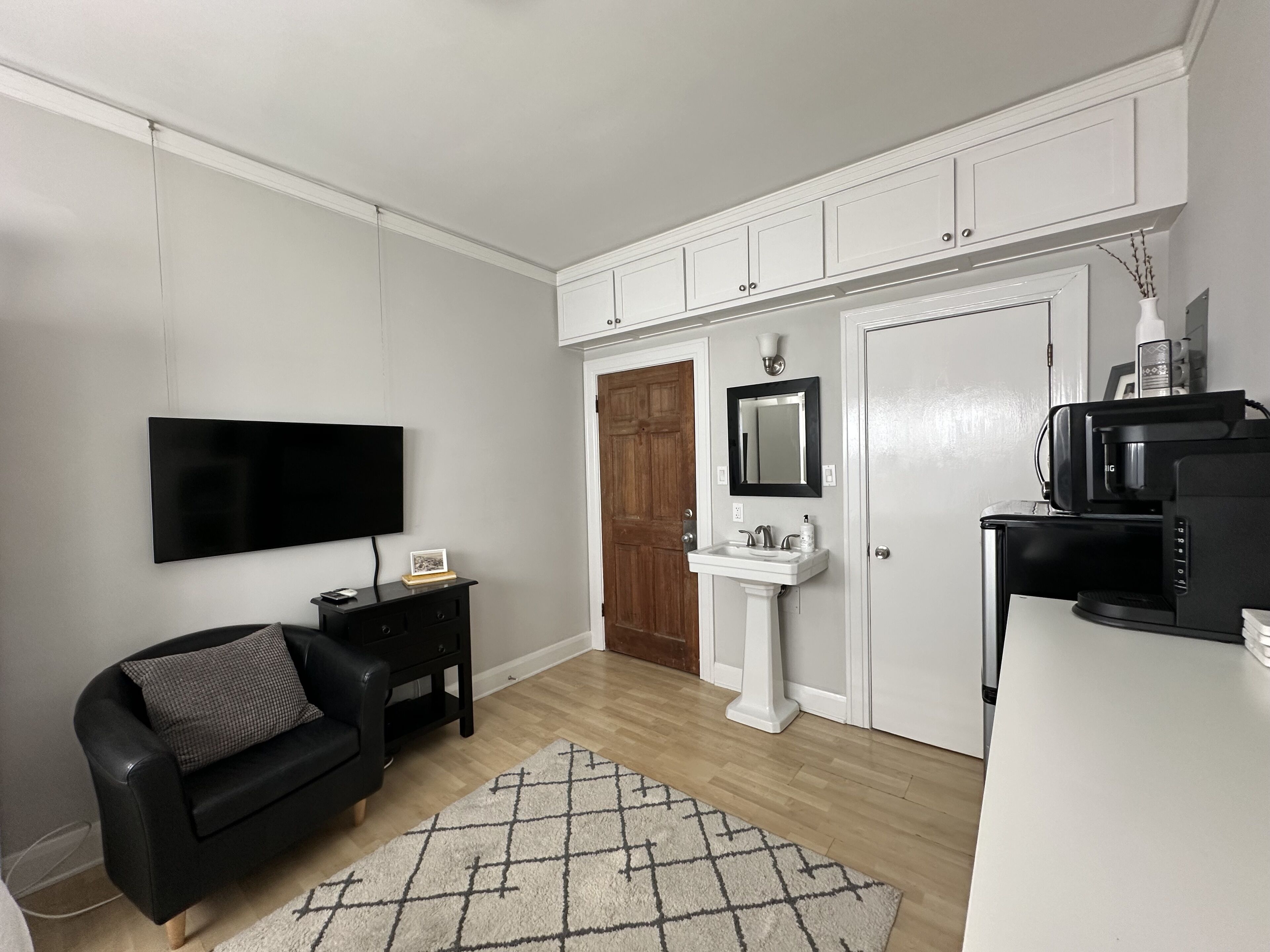 Een moderne woonkamer met een flatscreen televisie, een zwarte leren bank, een witte zuilwastafel en een koffiezetapparaat.