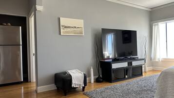 A modern living room with a large flat-screen TV, a black ottoman, a grey rug, and a wooden floor.