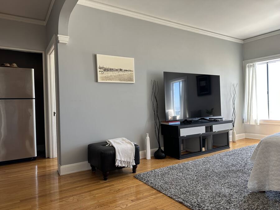 A modern living room with a large flat-screen TV, a black ottoman, a grey rug, and a wooden floor.