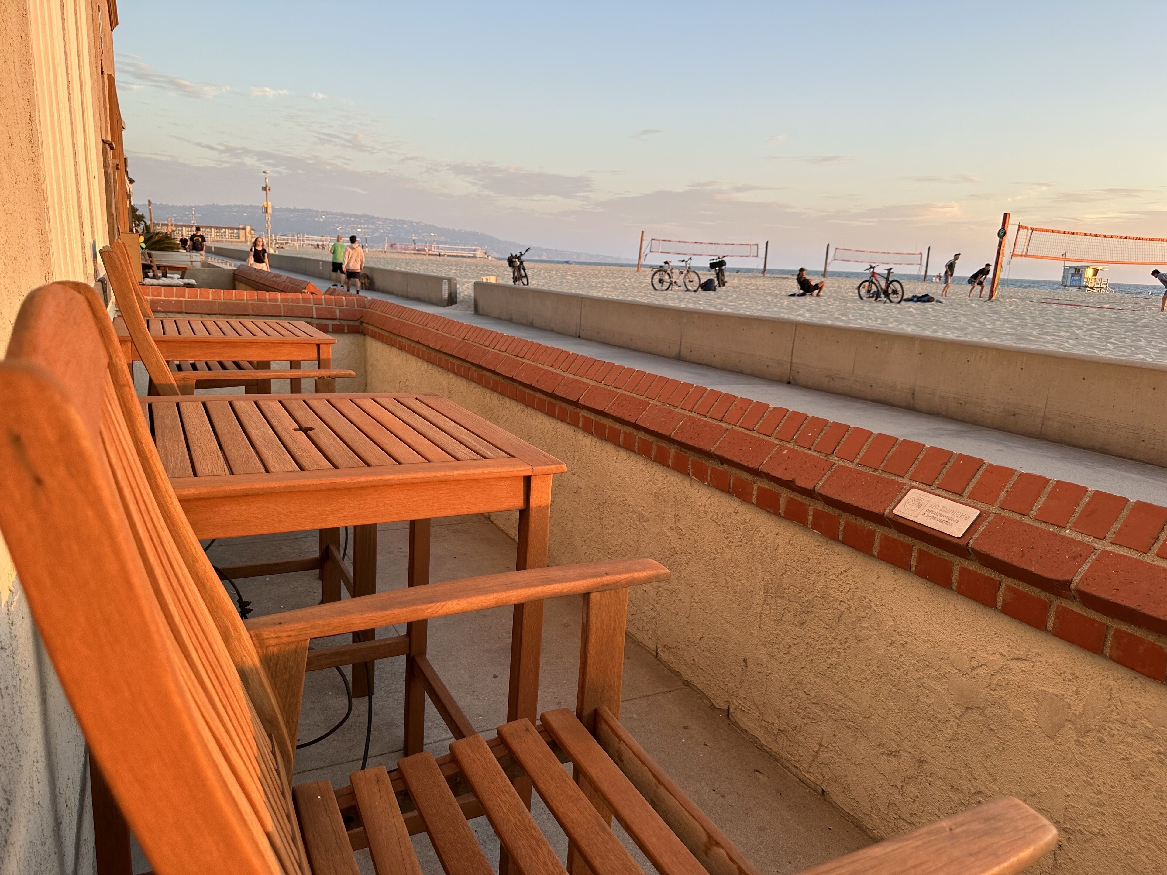 A beachside seating area with wooden tables and benches, a brick wall, and a volleyball net.