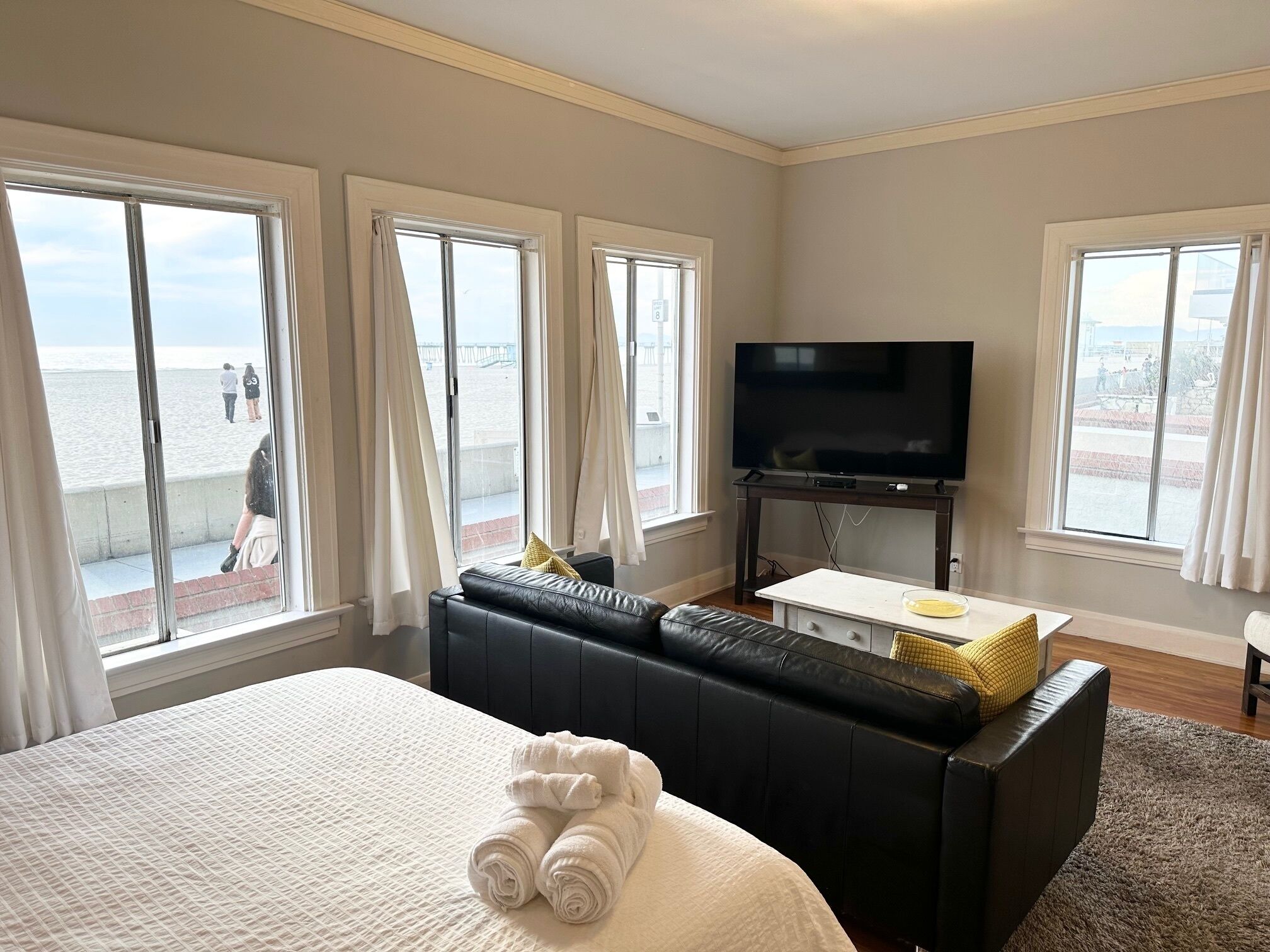 A hotel room with a large bed, a black sofa, a flat-screen TV, and a balcony with a view of the beach.