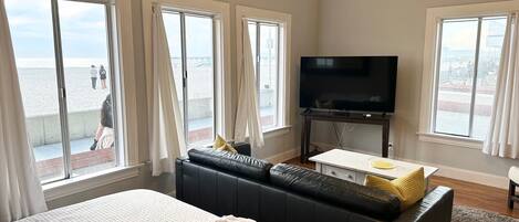 A hotel room with a large bed, a black sofa, a flat-screen TV, and a balcony with a view of the beach.