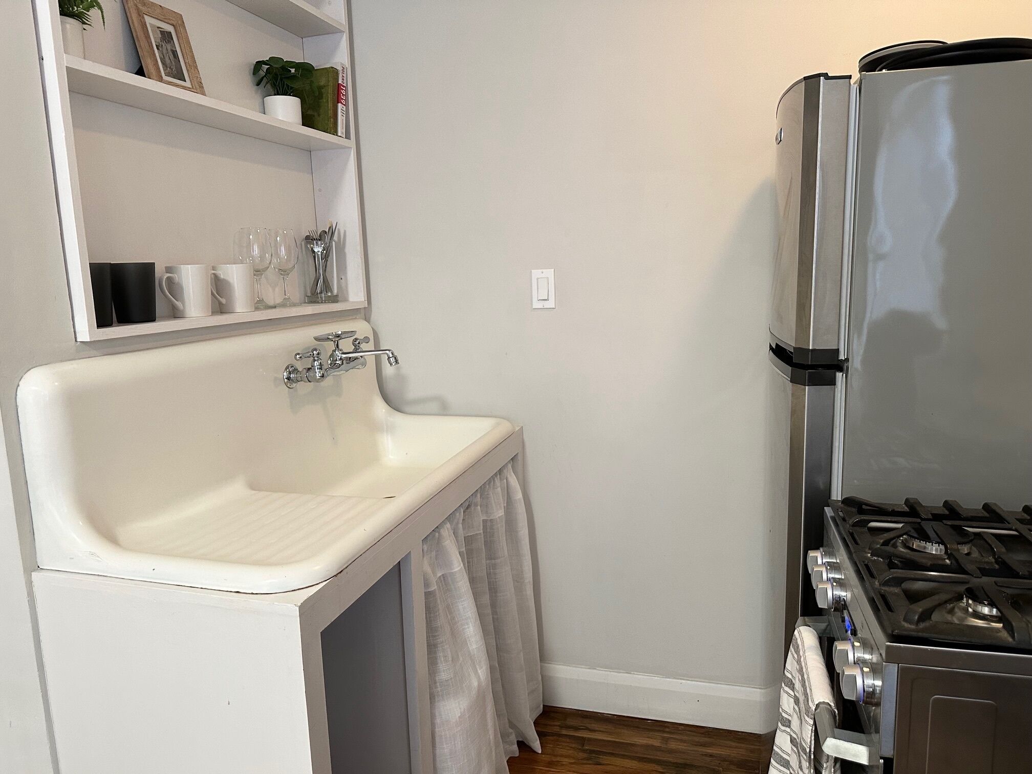 A compact kitchen with a white sink, a stove, and a shelf above the sink holding cups and a plant.