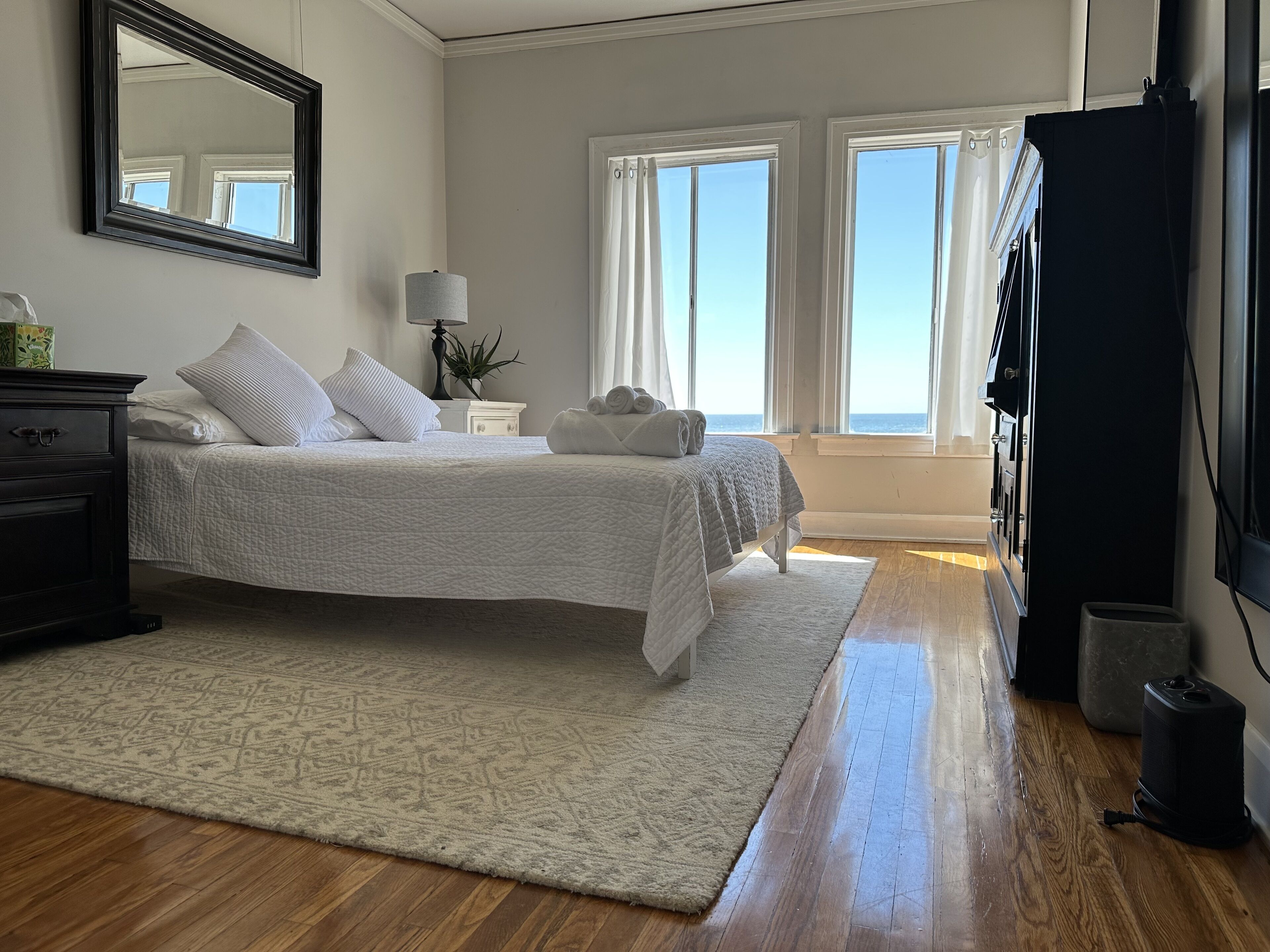 A bedroom with a bed, a dresser, a lamp, a TV, and a view of the ocean through large windows.