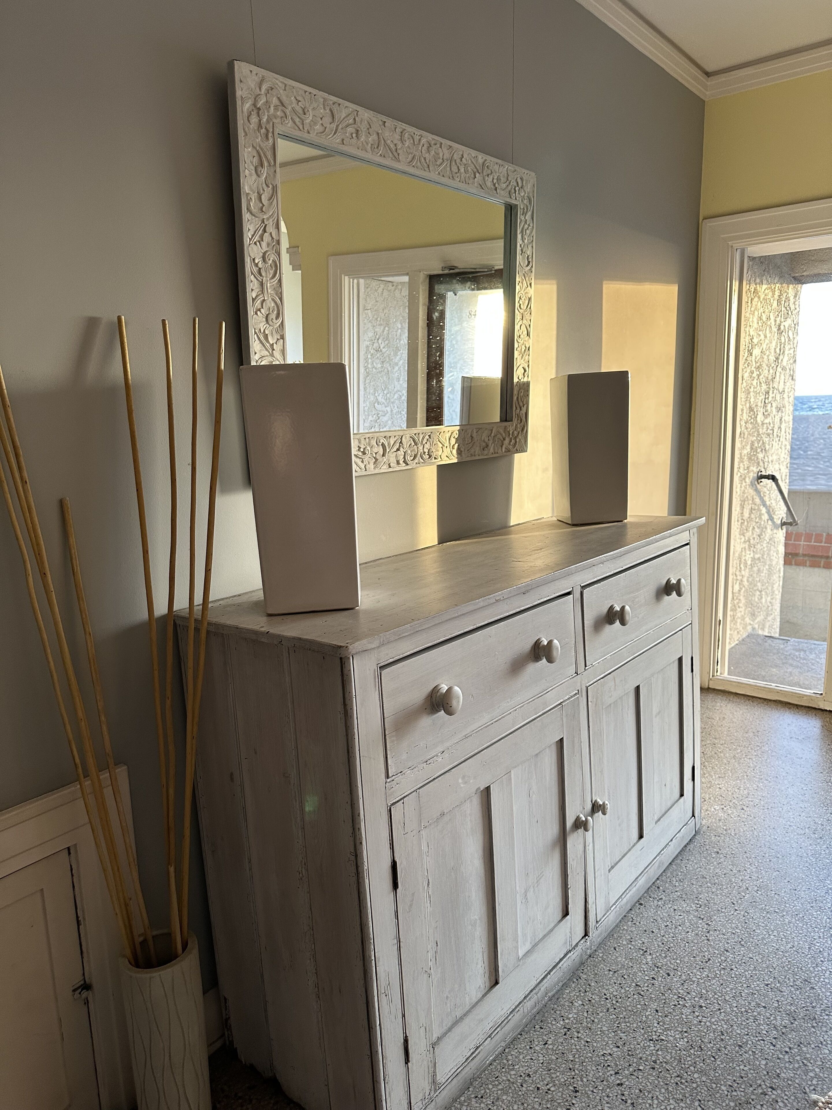 A white, distressed wooden dresser with a mirror above it, a vase with sticks, and a view of the sea through a glass door.