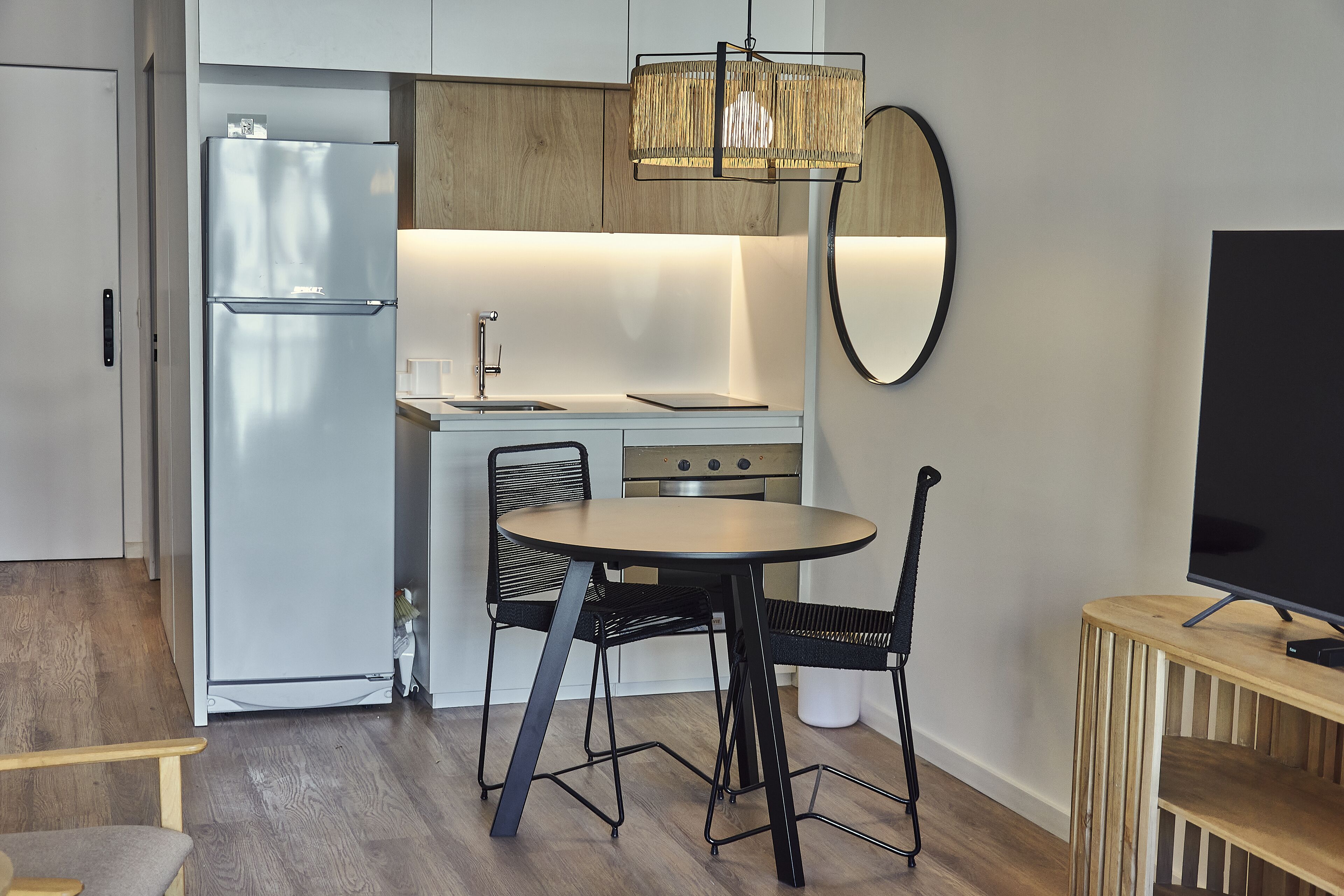 Una cocina compacta con refrigerador, pileta y estufa, una mesa redonda con sillas, y un televisor sobre un mueble de madera.