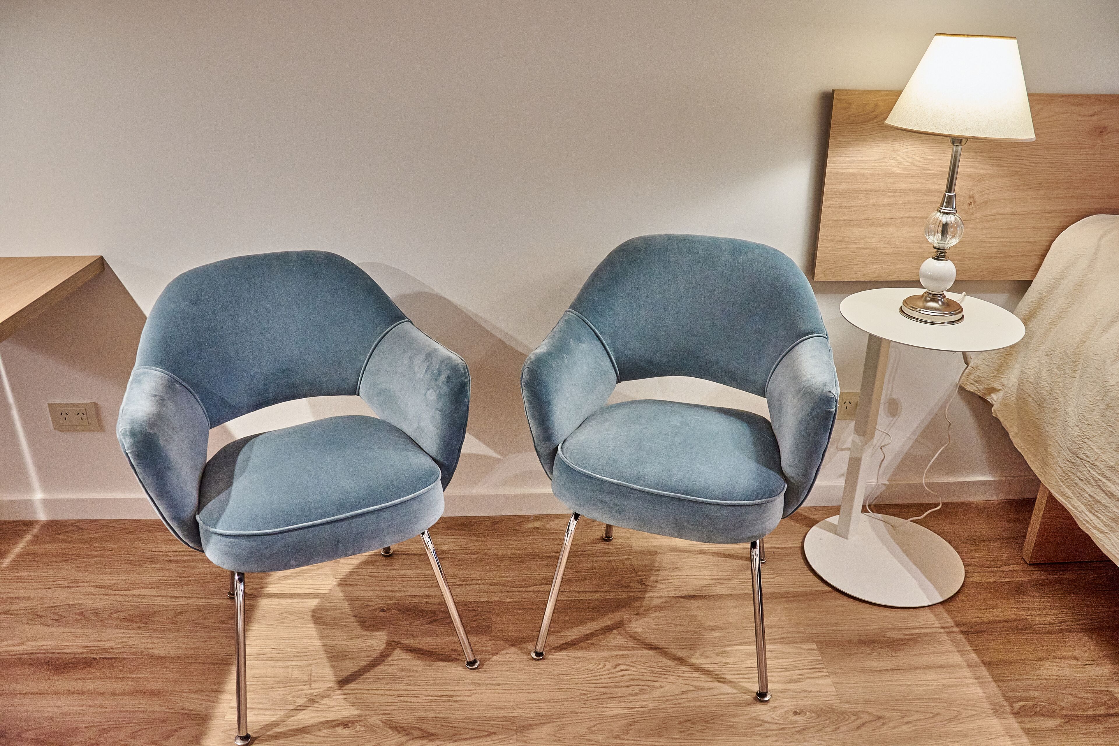 Two blue armchairs with metal legs, a white floor lamp, and a wooden headboard in a hotel room.