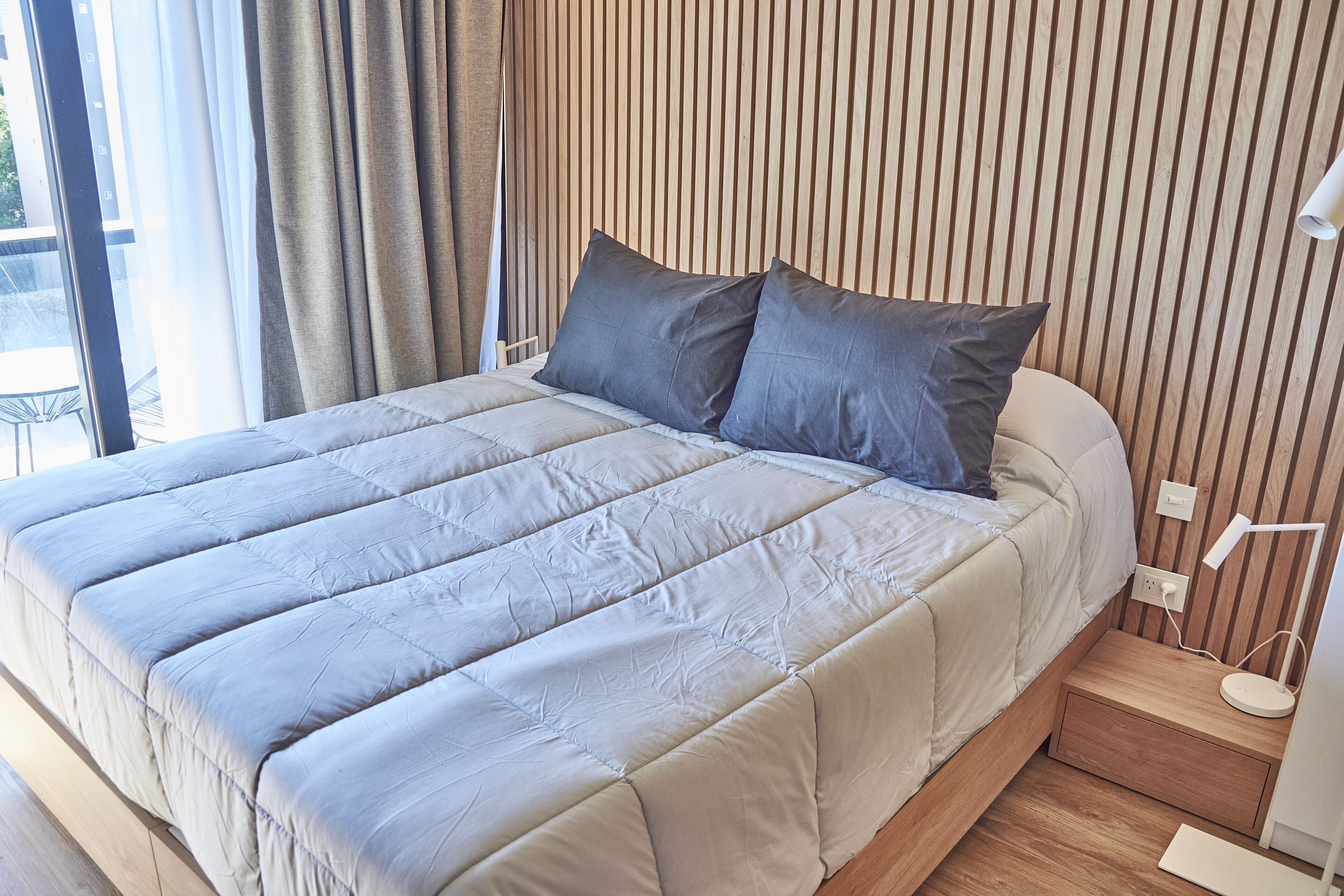 A neatly made bed with a light-colored duvet and two pillows, a wooden slatted headboard, a bedside table with a lamp, and a power outlet.