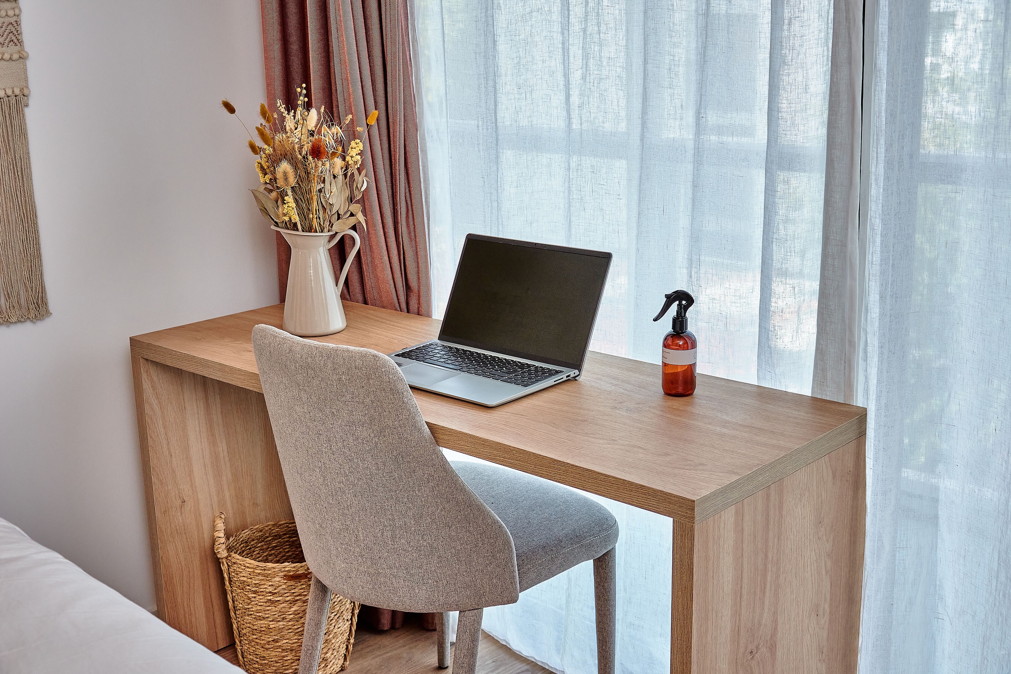 Een werkplek met een laptop, een vaas met gedroogde bloemen en een fles handdesinfectiemiddel op een houten bureau.