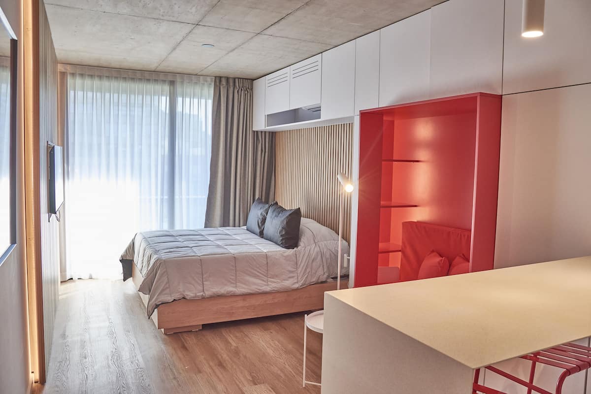 A modern bedroom with a bed, a desk, and a red accent wall.