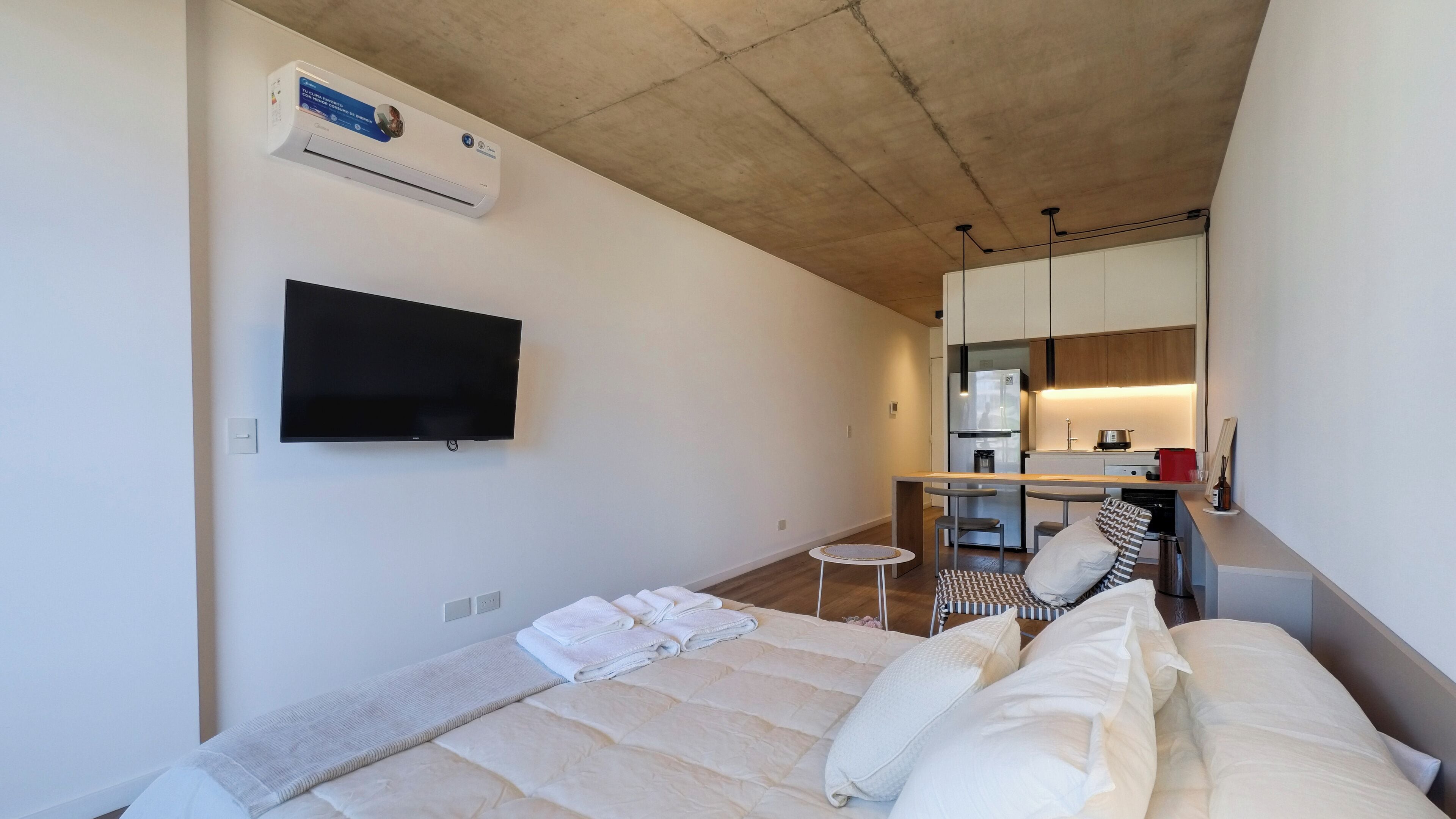 A modern hotel room with a bed, a flat-screen TV, a small round table, and a kitchenette in the background.